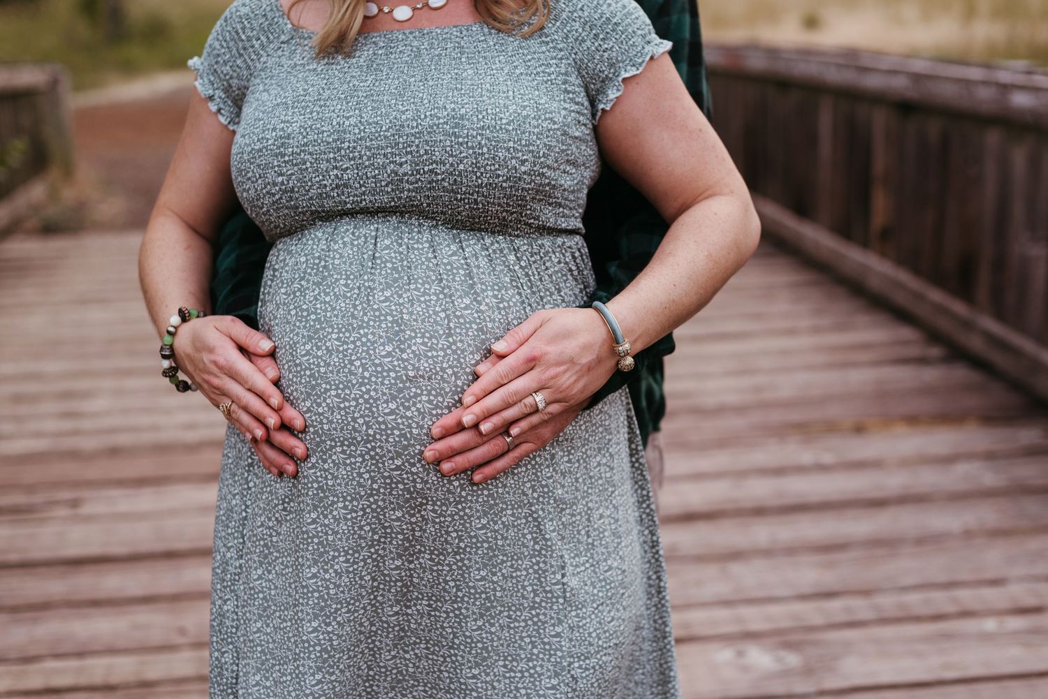 Close-up of pregnant belly with hands resting on gray smocked dress on wooden boardwalk.