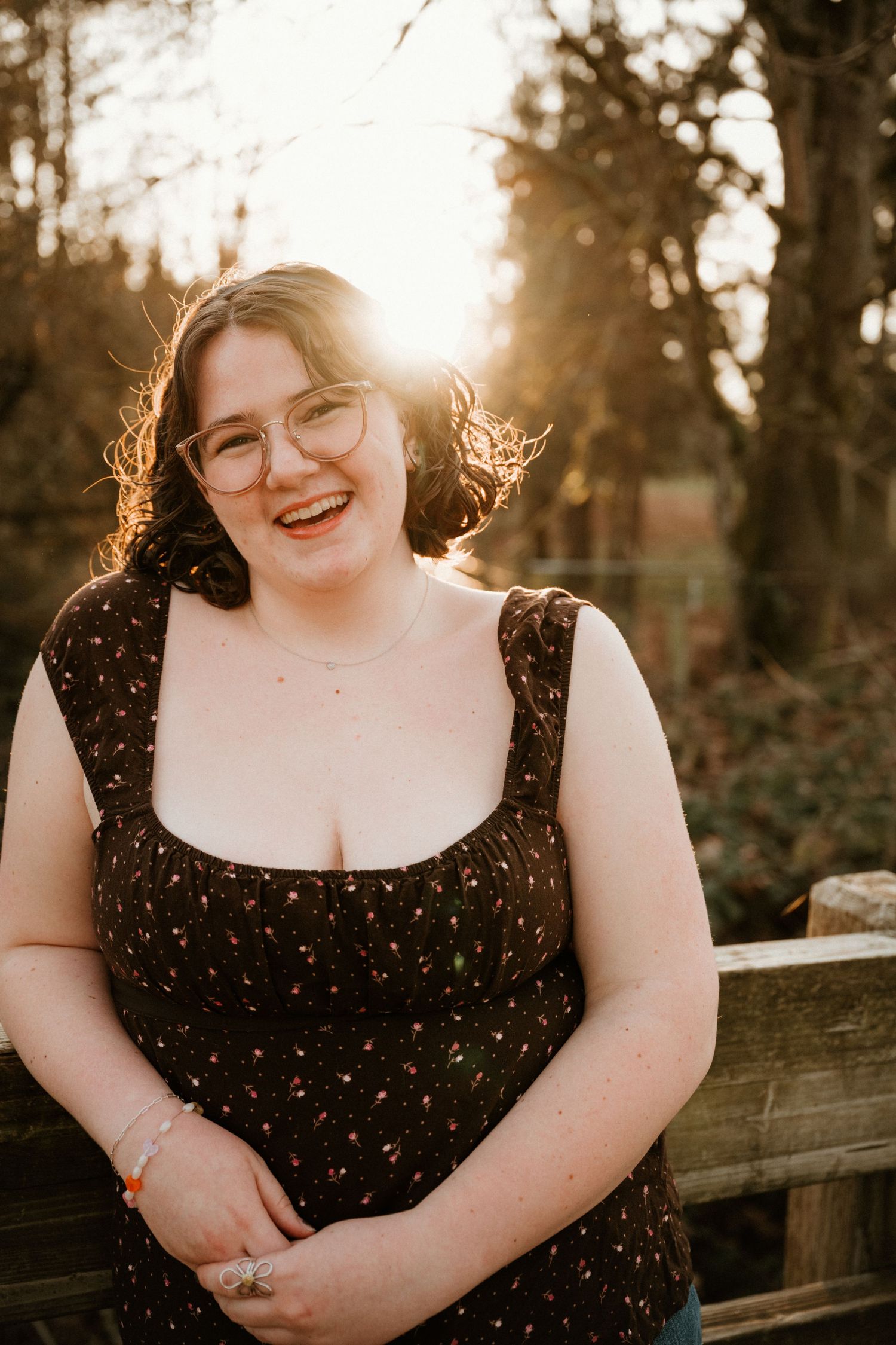A person in a black floral dress smiles warmly during a sunset photoshoot against a wooden fence.