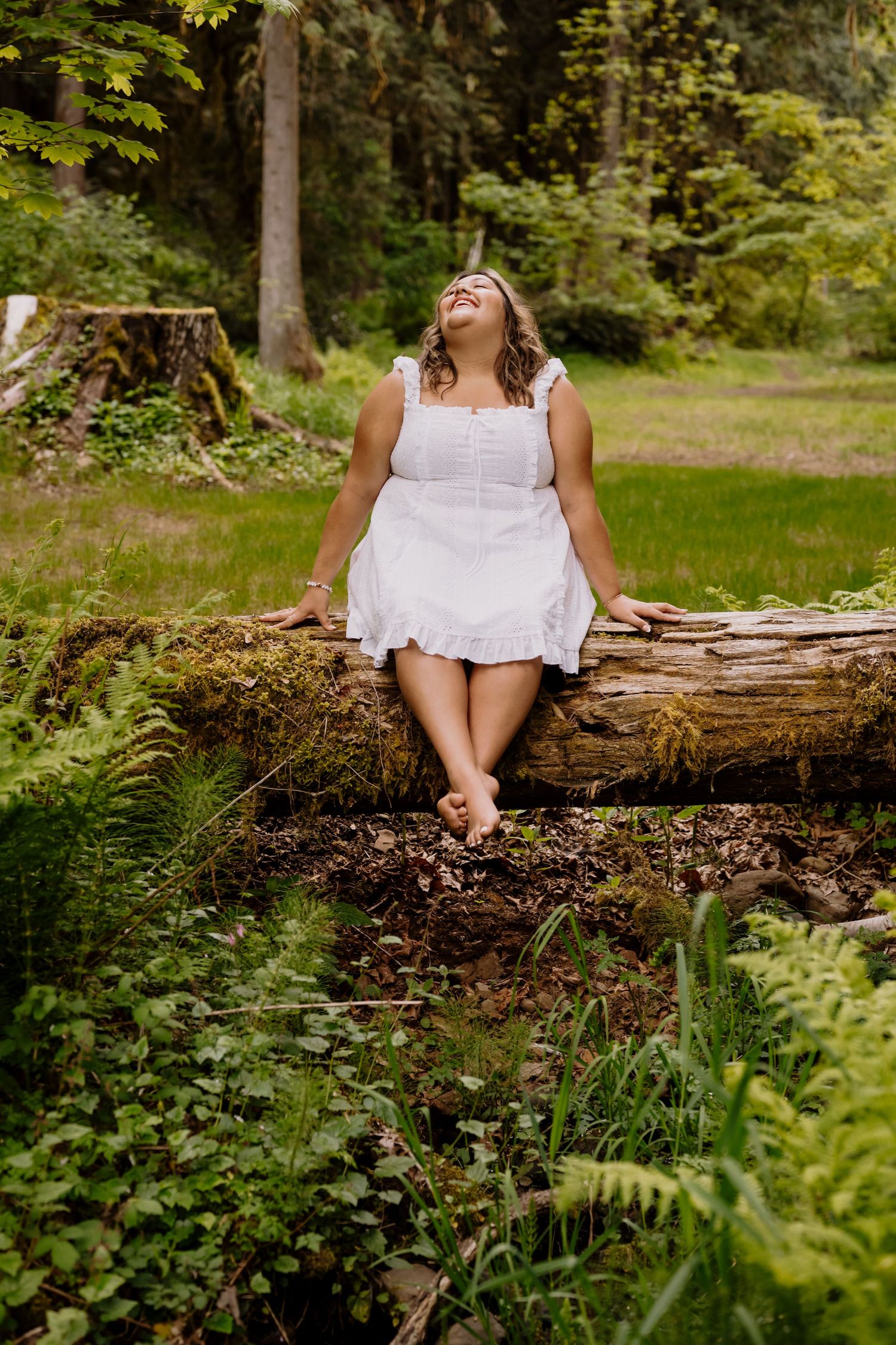 Woman in white dress sitting on mossy log, laughing in serene forest setting.