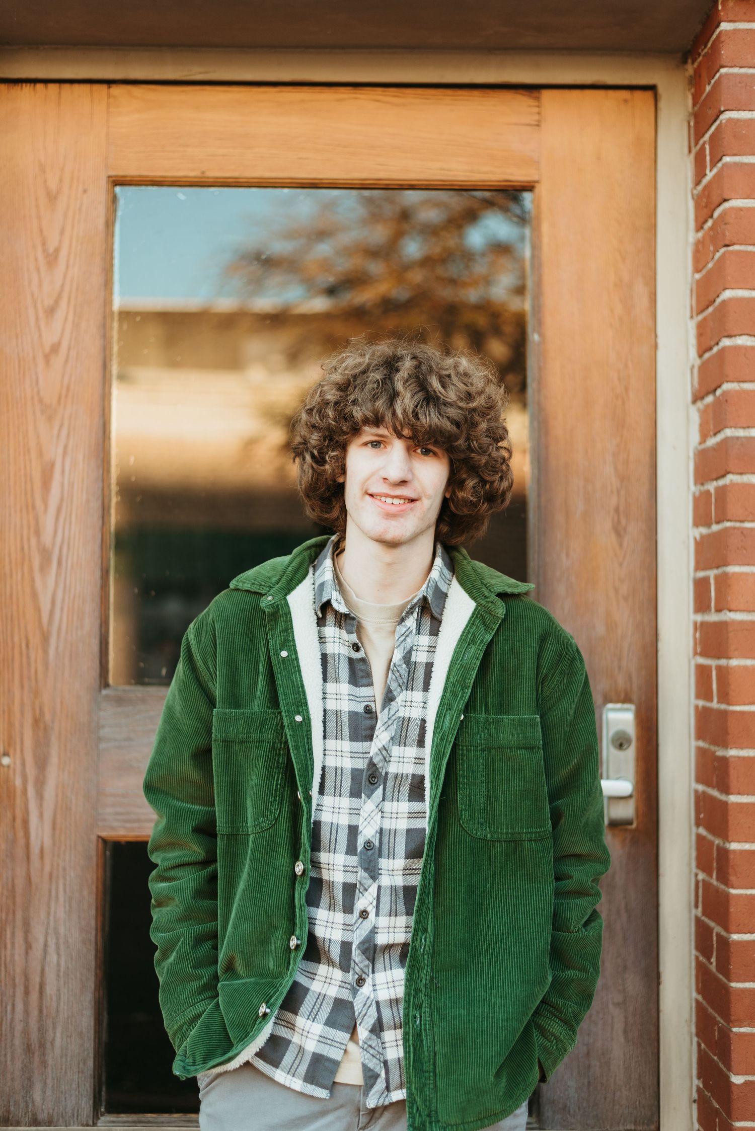Young person in green corduroy jacket and plaid shirt stands by wooden door.