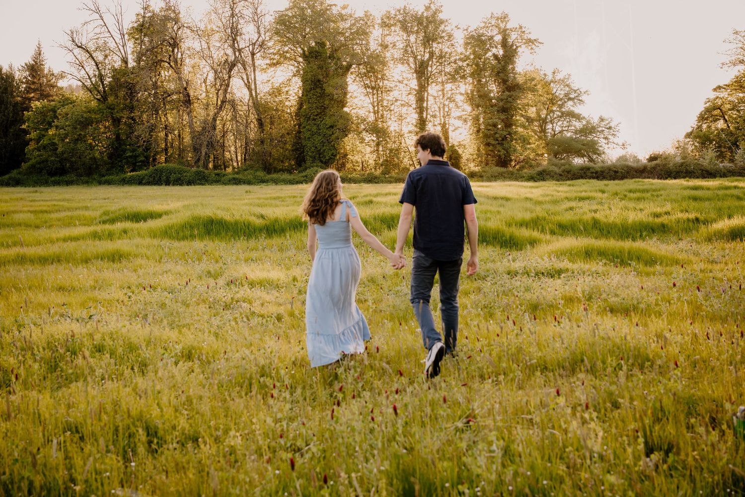 Couple holding hands walking through a lush green field at golden hour with trees in the background.