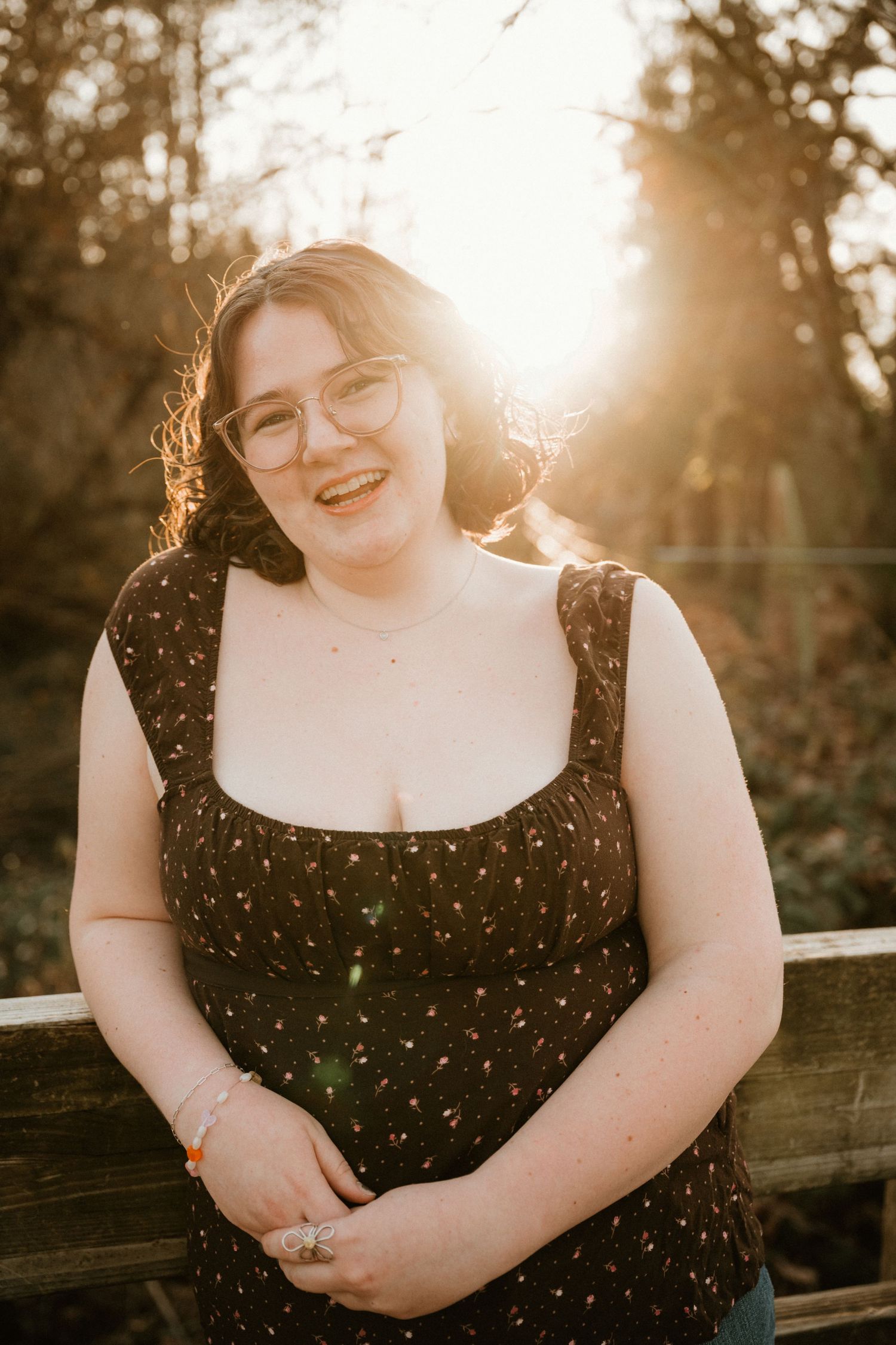 Young woman in black floral top and glasses smiling outdoors at golden hour with warm sunlight behind her.