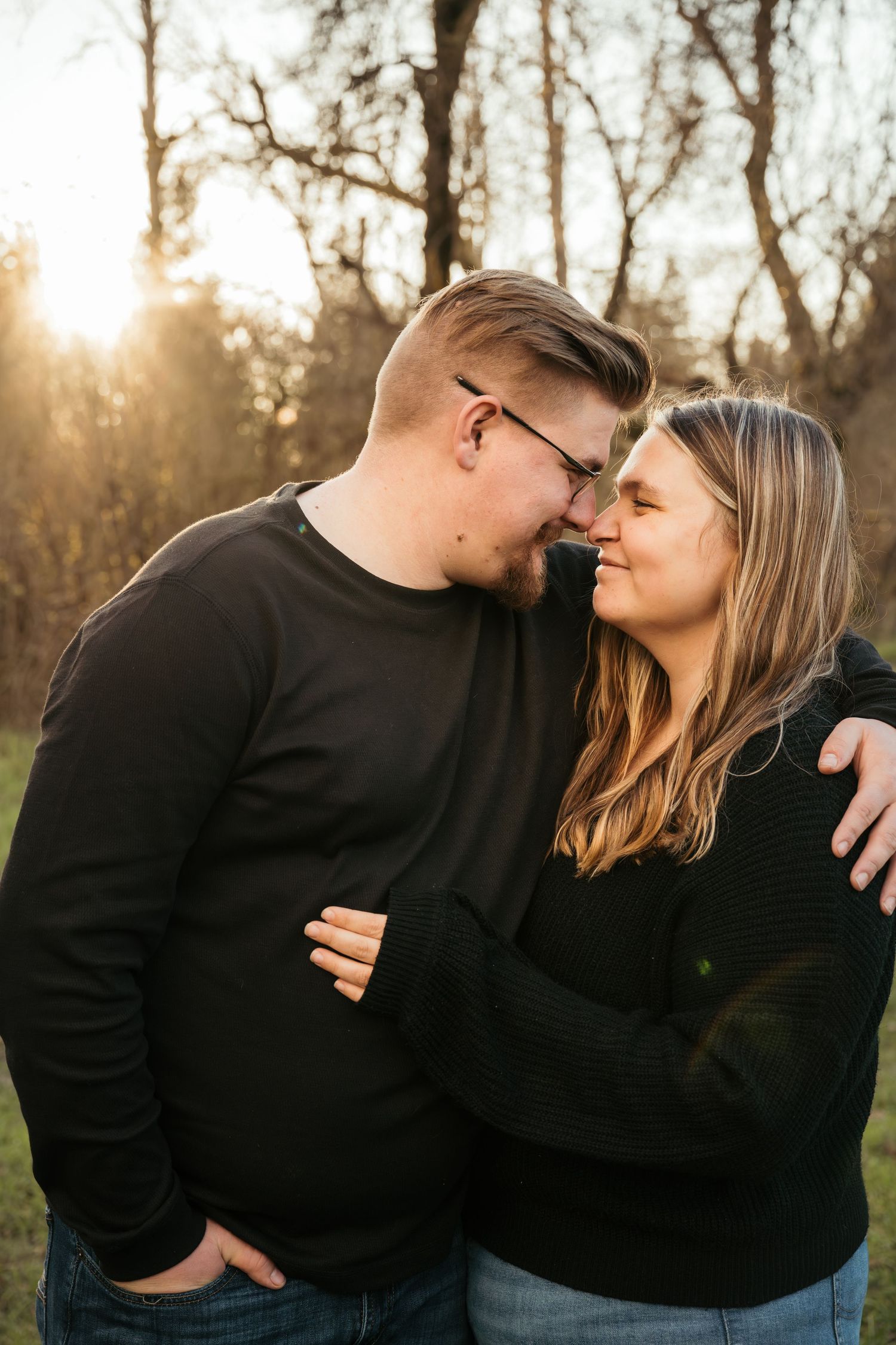 A romantic couple in matching black sweaters share intimate moments in a wooded area at sunset.