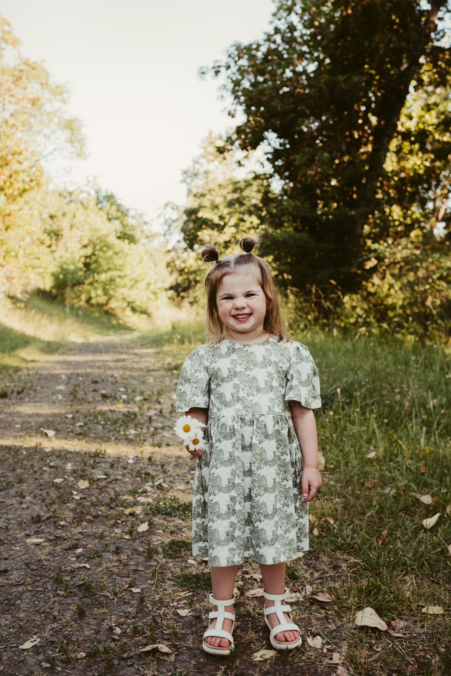Little girl in a floral dress holds a daisy while standing on a nature trail.
