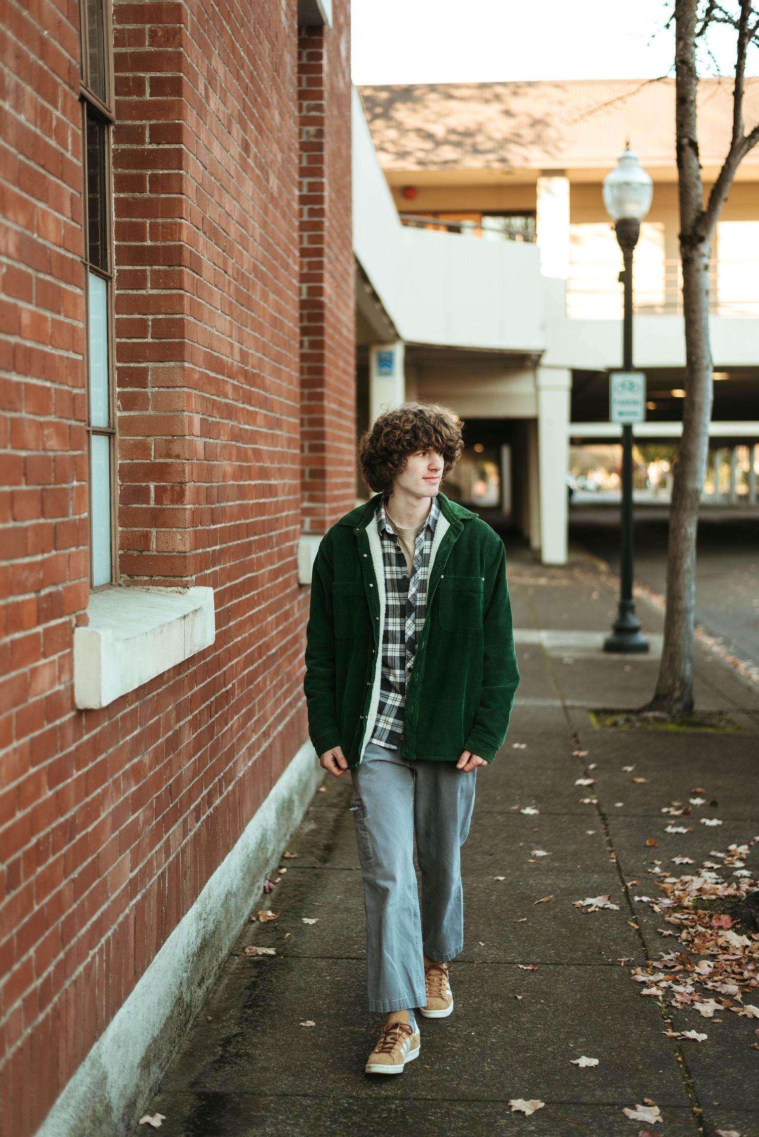 Person in green jacket and plaid shirt walks along brick wall on urban sidewalk.