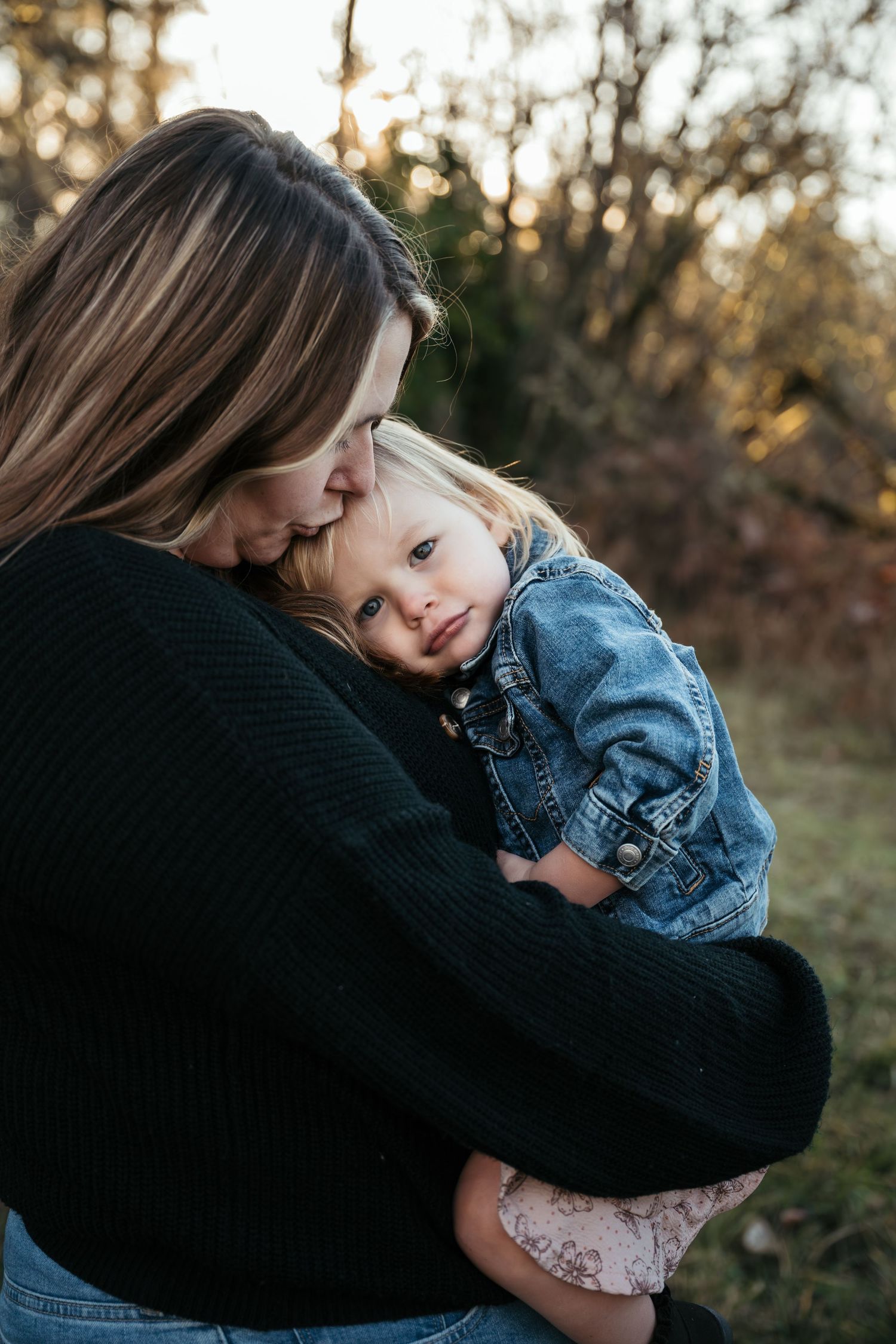 A mother tenderly holds and kisses her toddler daughter outdoors in soft golden light.
