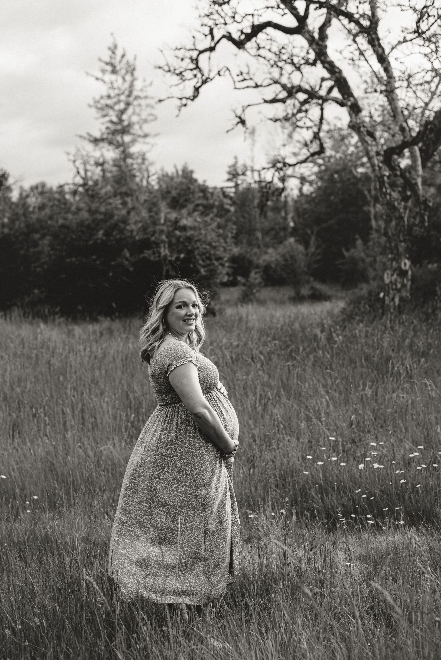 Black and white portrait of expectant mother in flowing dress standing in meadow.