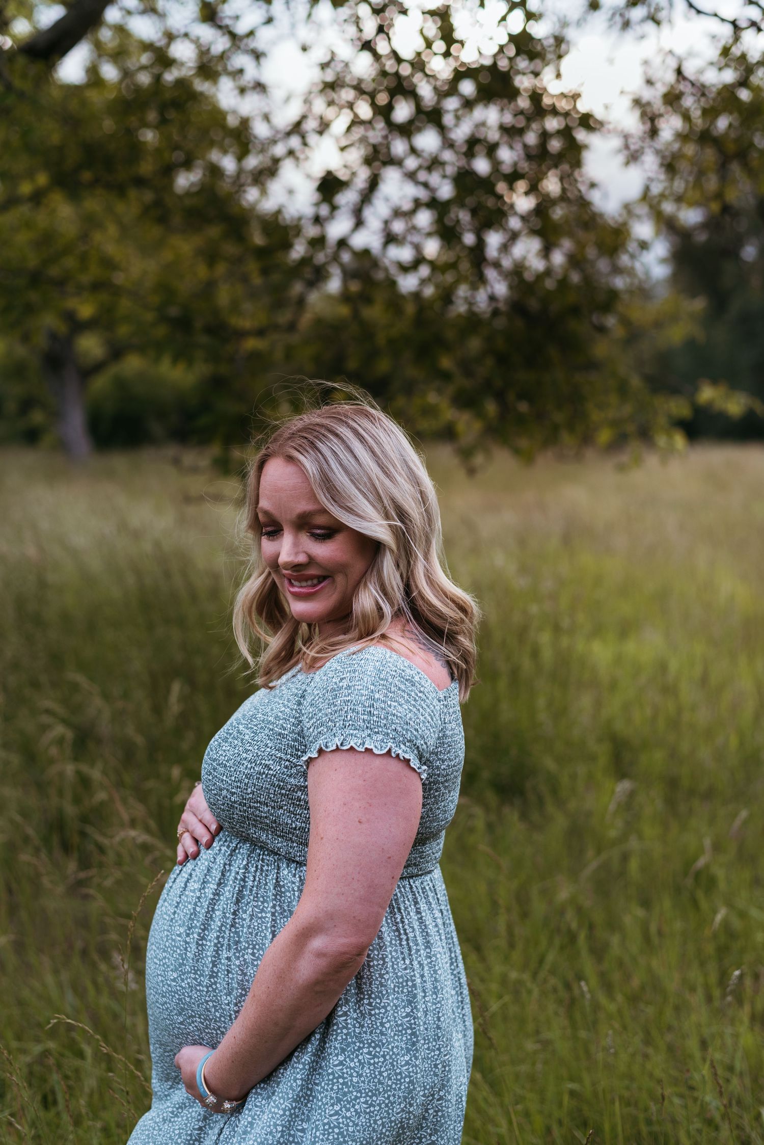 Expectant mother in a blue floral dress poses in a serene meadow during golden hour.