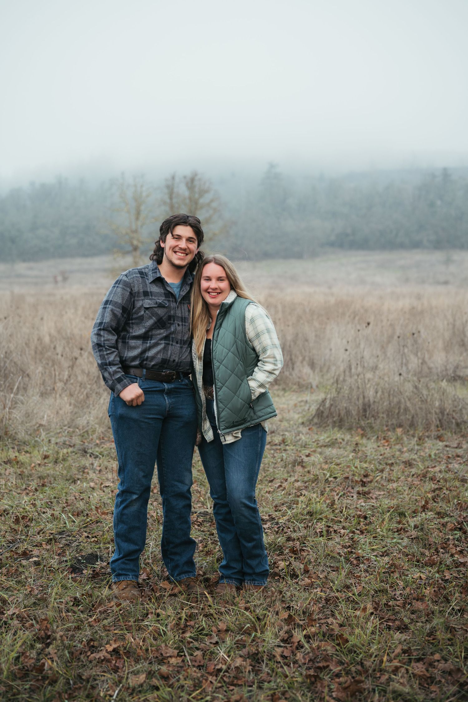 Two people wearing casual outdoor attire pose together in a foggy field during autumn.