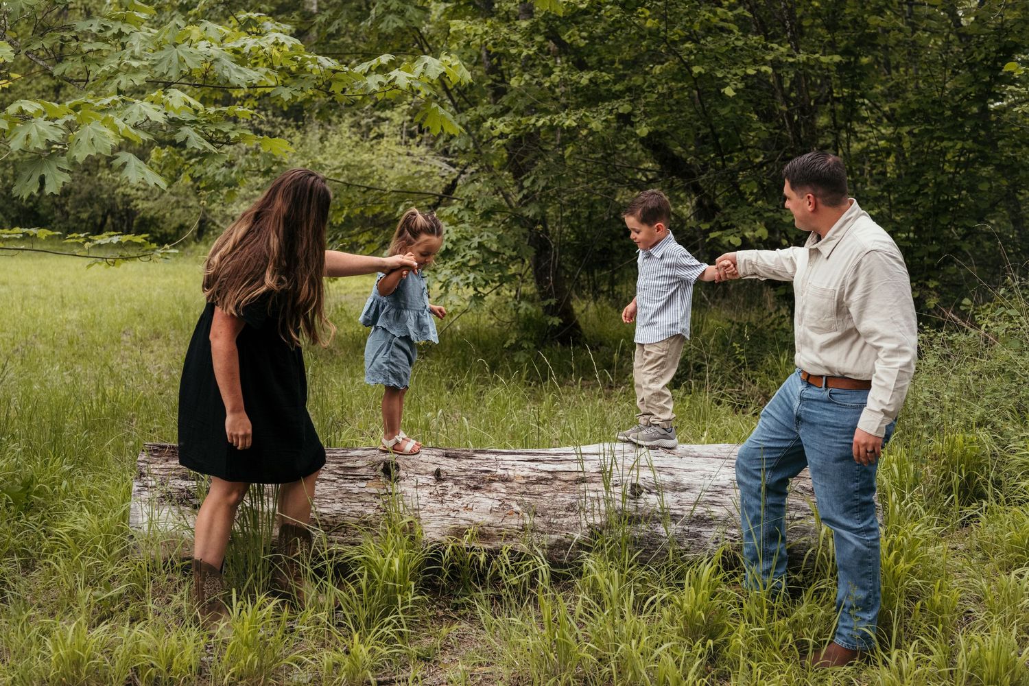 Family of four playing on a fallen log in a lush green meadow surrounded by trees.