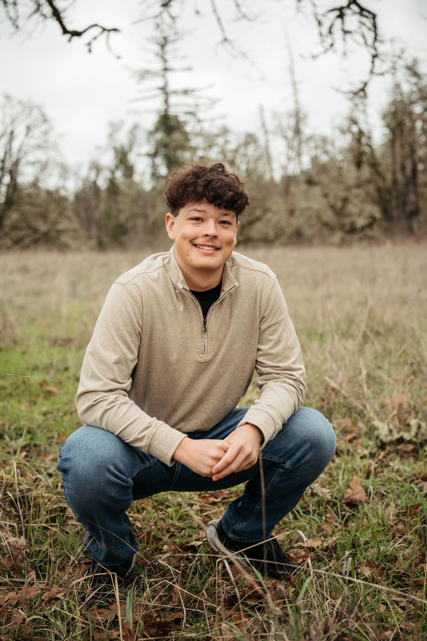Young man crouching in a grassy field, smiling, wearing a beige quarter-zip sweater and jeans, with trees in background.