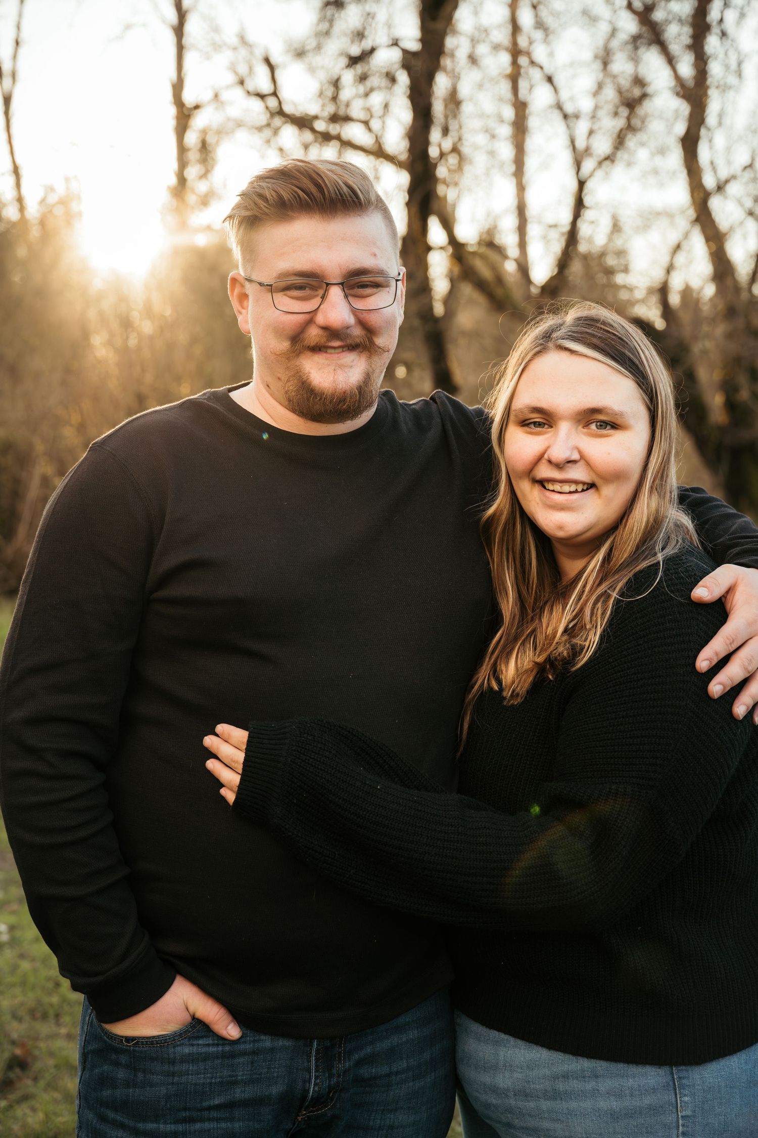 Couple wearing matching black sweaters embraces during outdoor sunset photoshoot in wooded setting.