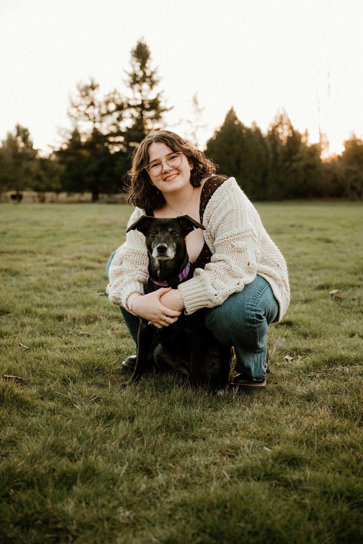A smiling young woman with glasses crouches on green grass hugging a small black dog at golden hour.