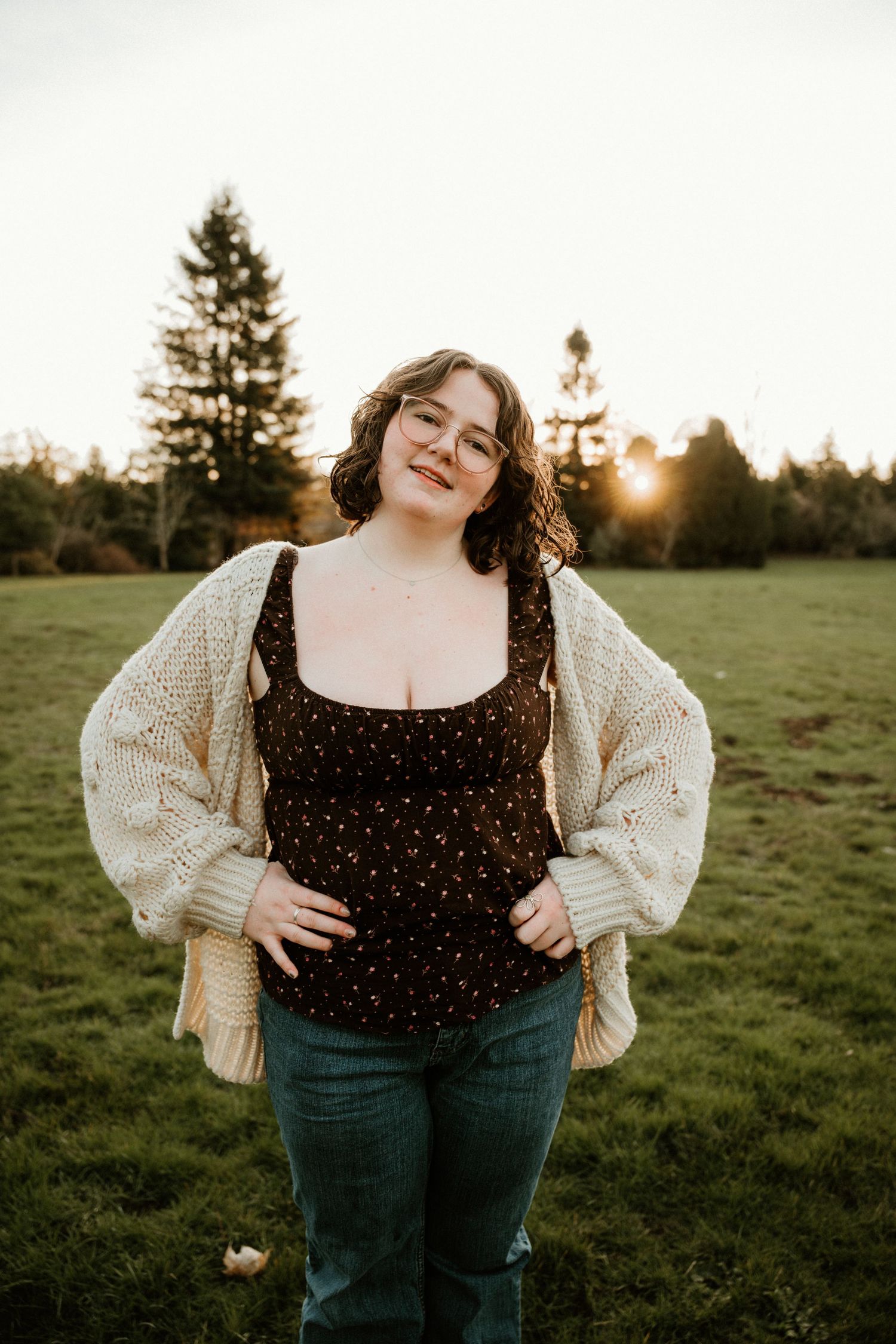 A person in a polka dot top and jeans poses in a grassy field at sunset.