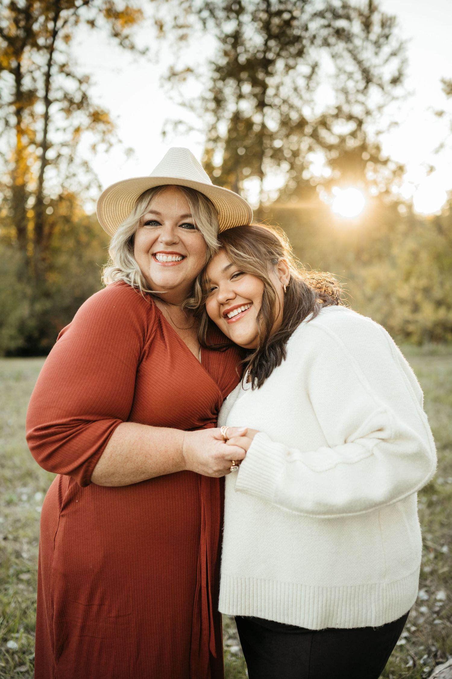 Two women smiling and embracing outdoors at sunset, one in a rust dress with hat, other in white sweater.