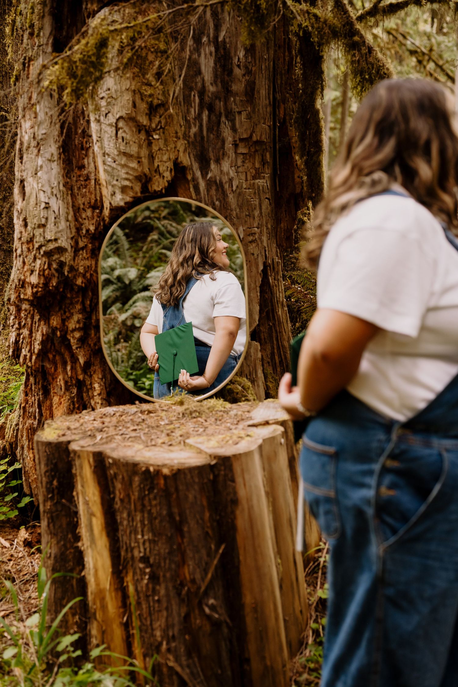 Woman in white tee and green overalls views her reflection in a round mirror propped against a mossy forest tree.