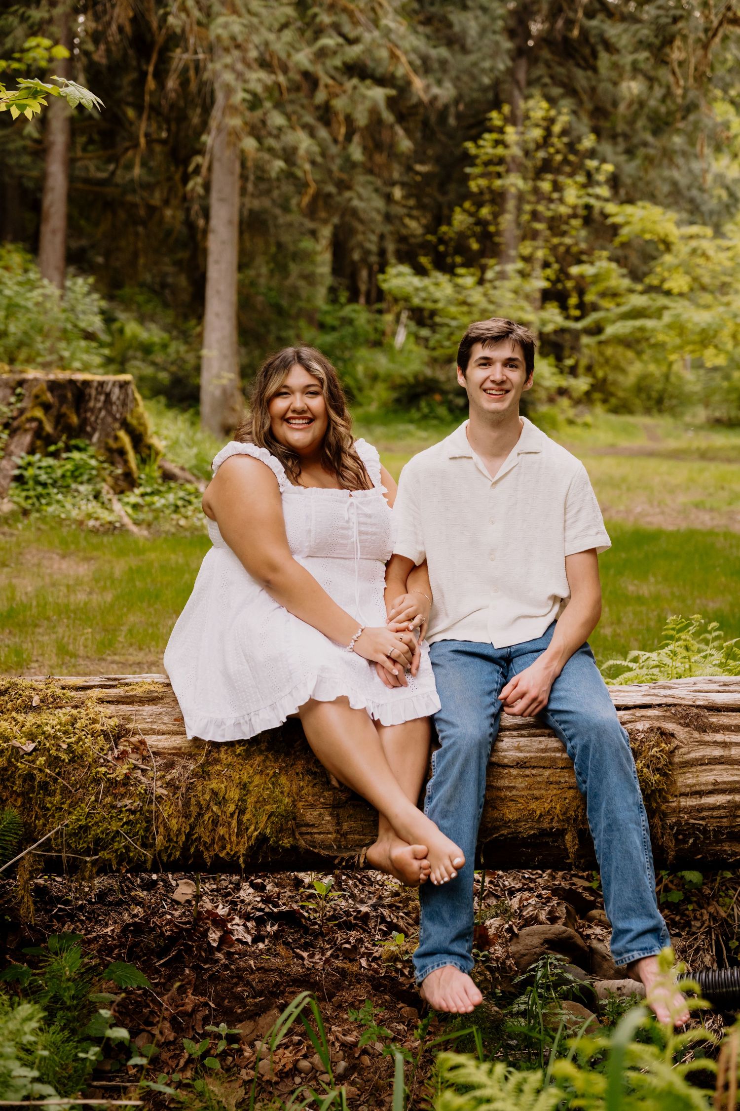 A smiling couple sits barefoot on a mossy log in a lush green forest, wearing white outfits and holding hands.