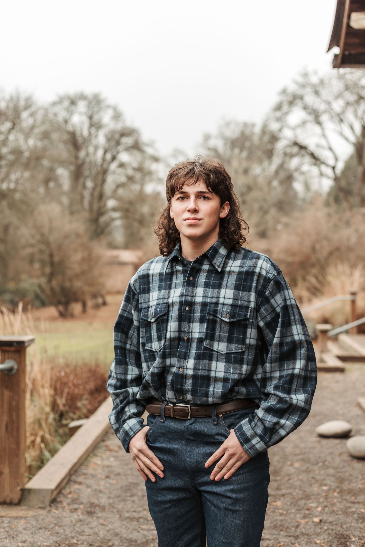 Person wearing plaid flannel shirt and jeans stands outdoors against natural backdrop with trees.