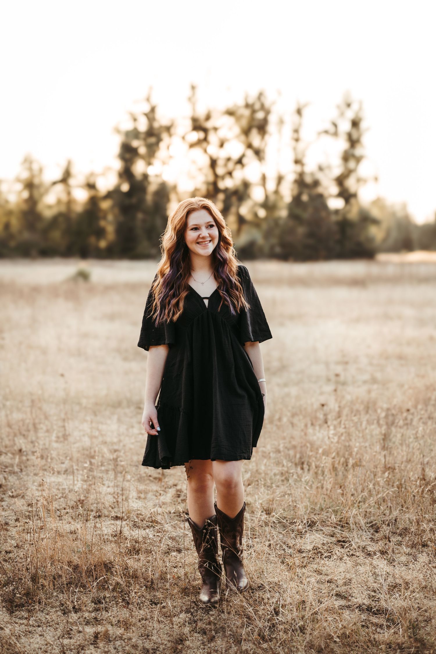 Woman in a black dress and boots poses in a golden field at sunset.
