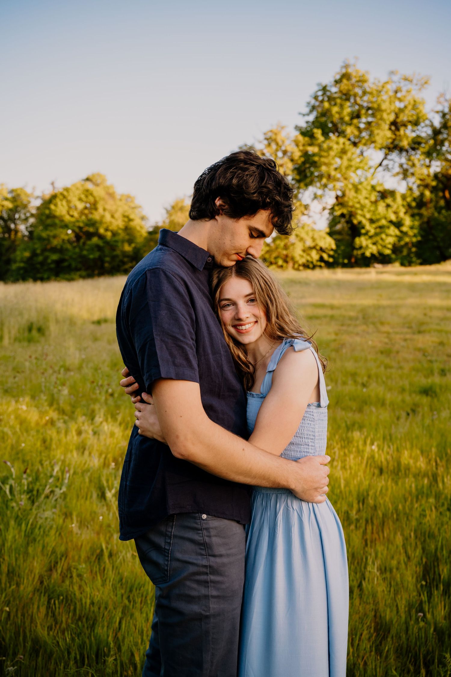 A couple embraces in a sunny green field, the woman in a blue dress smiling warmly at the camera.