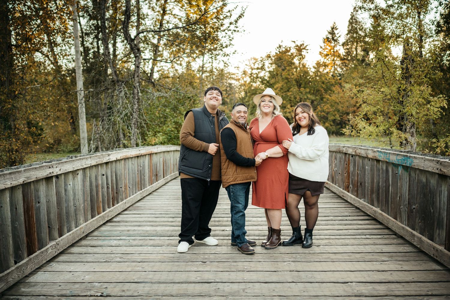 Four people smiling together on a wooden bridge surrounded by autumn trees in a warm family portrait.