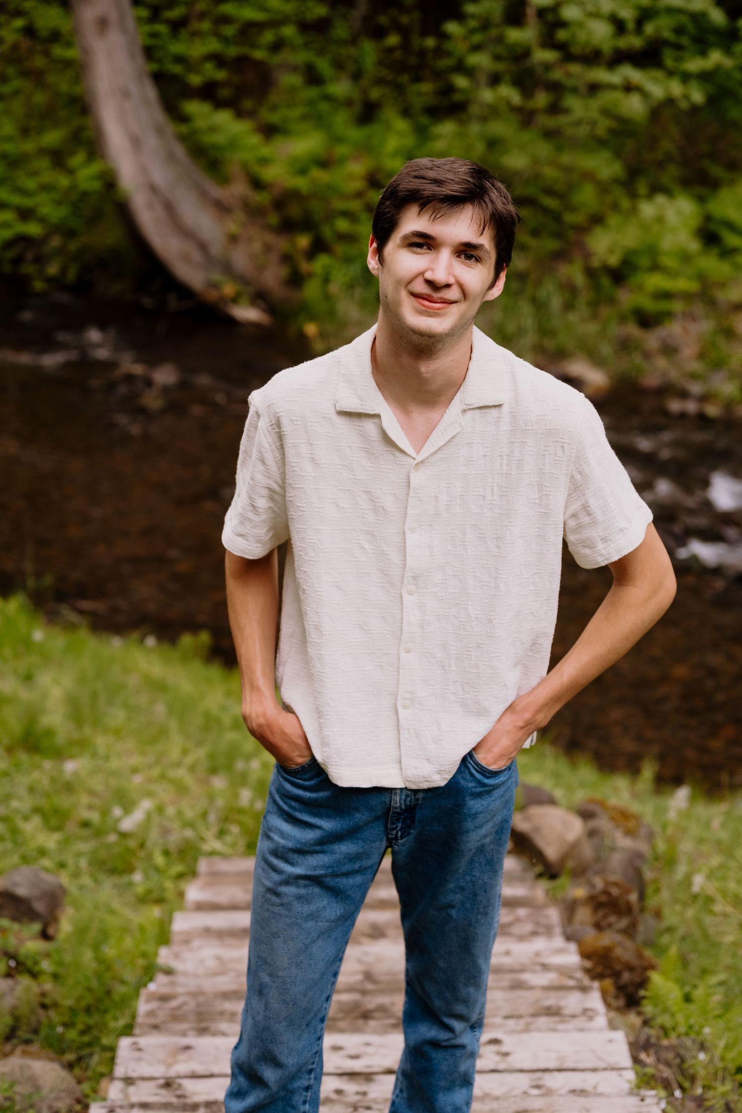 Young man in white linen shirt and jeans standing on wooden bridge by stream in lush green forest.