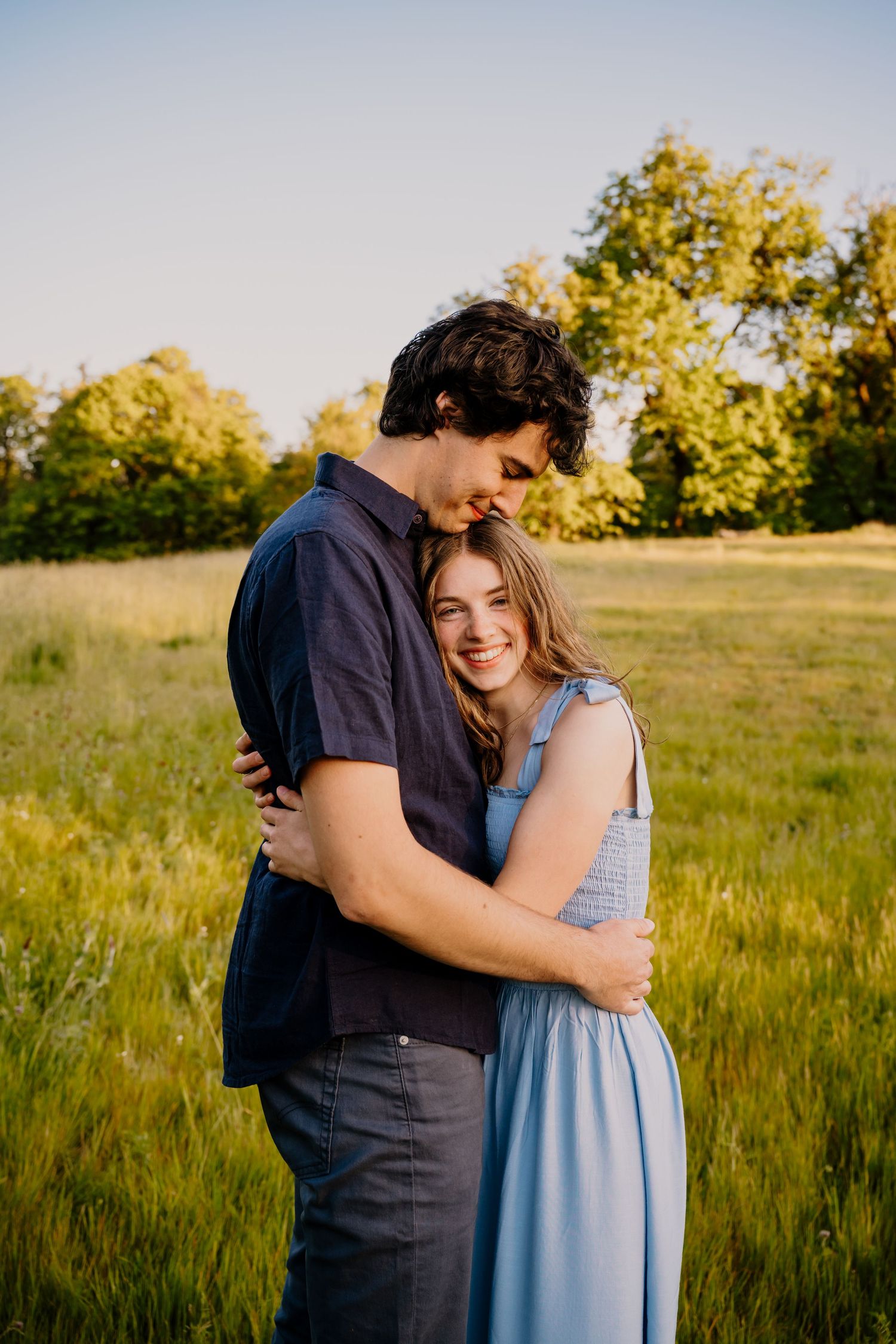 A young couple embraces in a sunlit green meadow, the woman in a blue dress smiling warmly as golden hour light glows behind them.