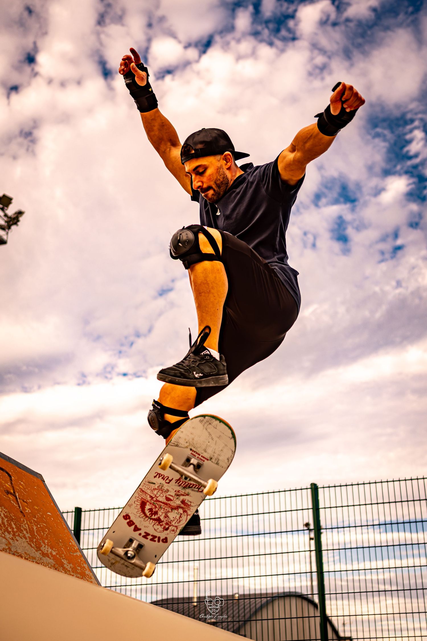 Skateboarder performs aerial trick against cloudy sky at skate park with fence in background.