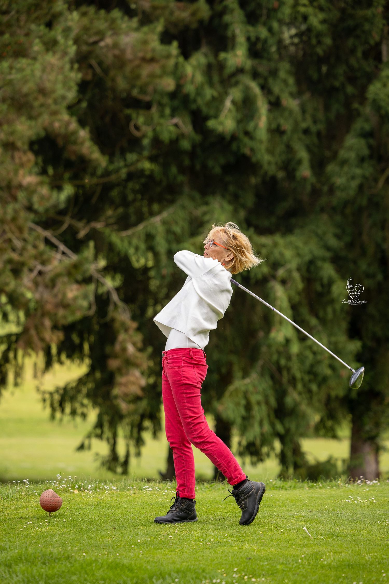 Young golfer in a white pullover and red pants takes a swing at a golf ball on a lush green course.
