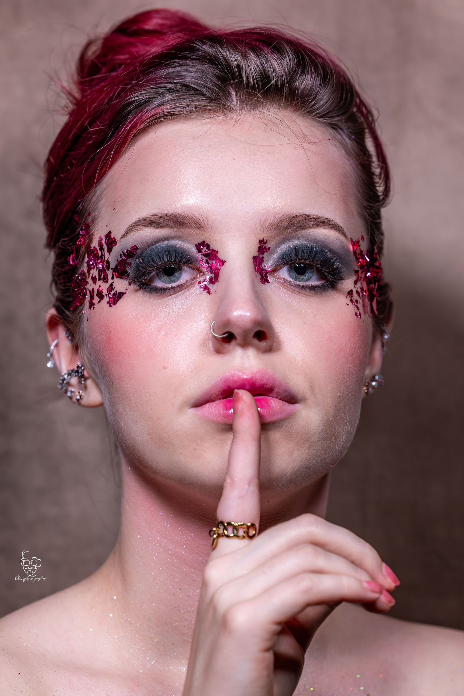 Creative beauty portrait with dramatic makeup featuring pink glitter eye art, burgundy hair, and shushing gesture.