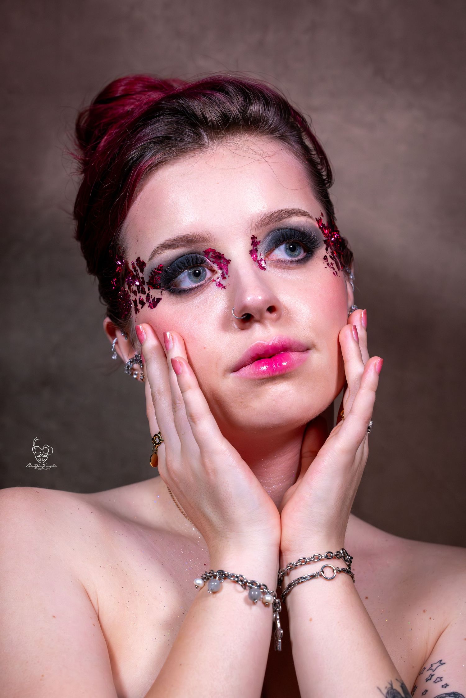 Artistic beauty portrait with dramatic pink makeup, intricate eye decorations, and delicate silver bracelets against dark background.