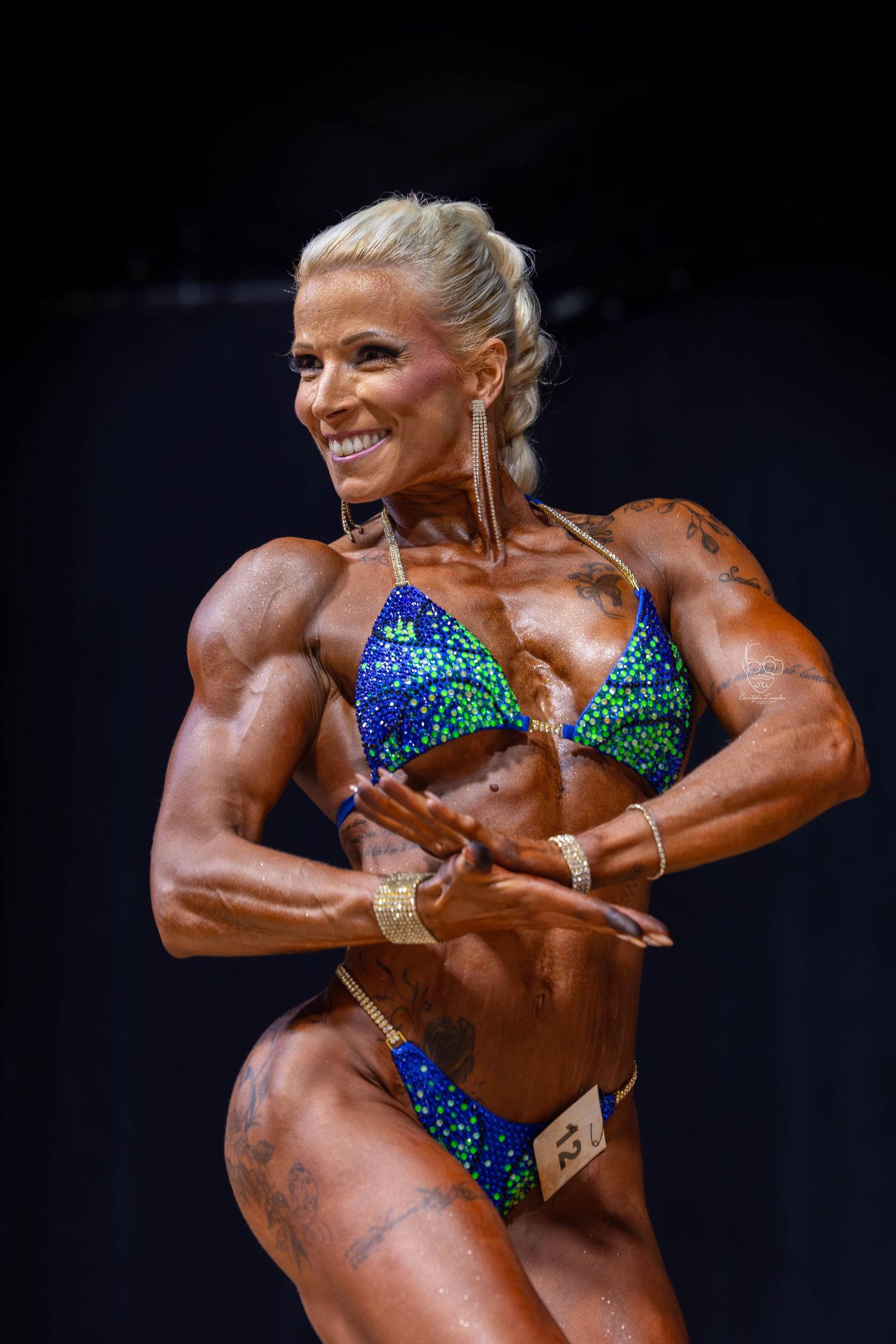 Bodybuilder in sparkly blue and green competition bikini strikes different poses on dark stage during competition.