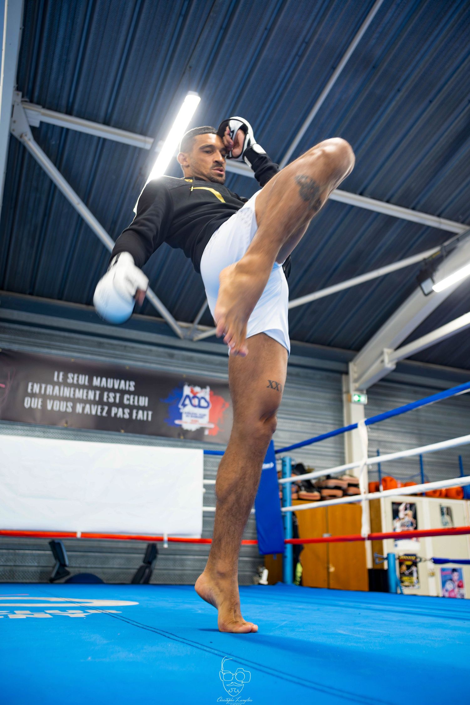 Martial artist executing a high kick during training in a boxing gym with blue mat and industrial ceiling.