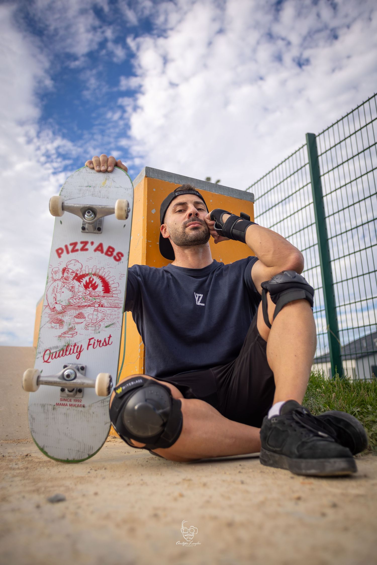 A skateboarder takes a break with their board and water bottle against a fence on a sunny day with cloudy blue skies.