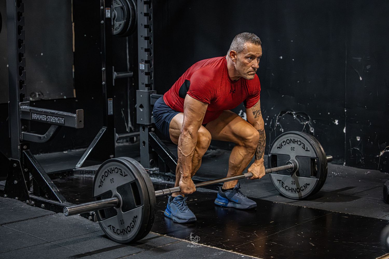 A person in a red shirt performs a deadlift exercise with a loaded barbell in a dark gym setting.