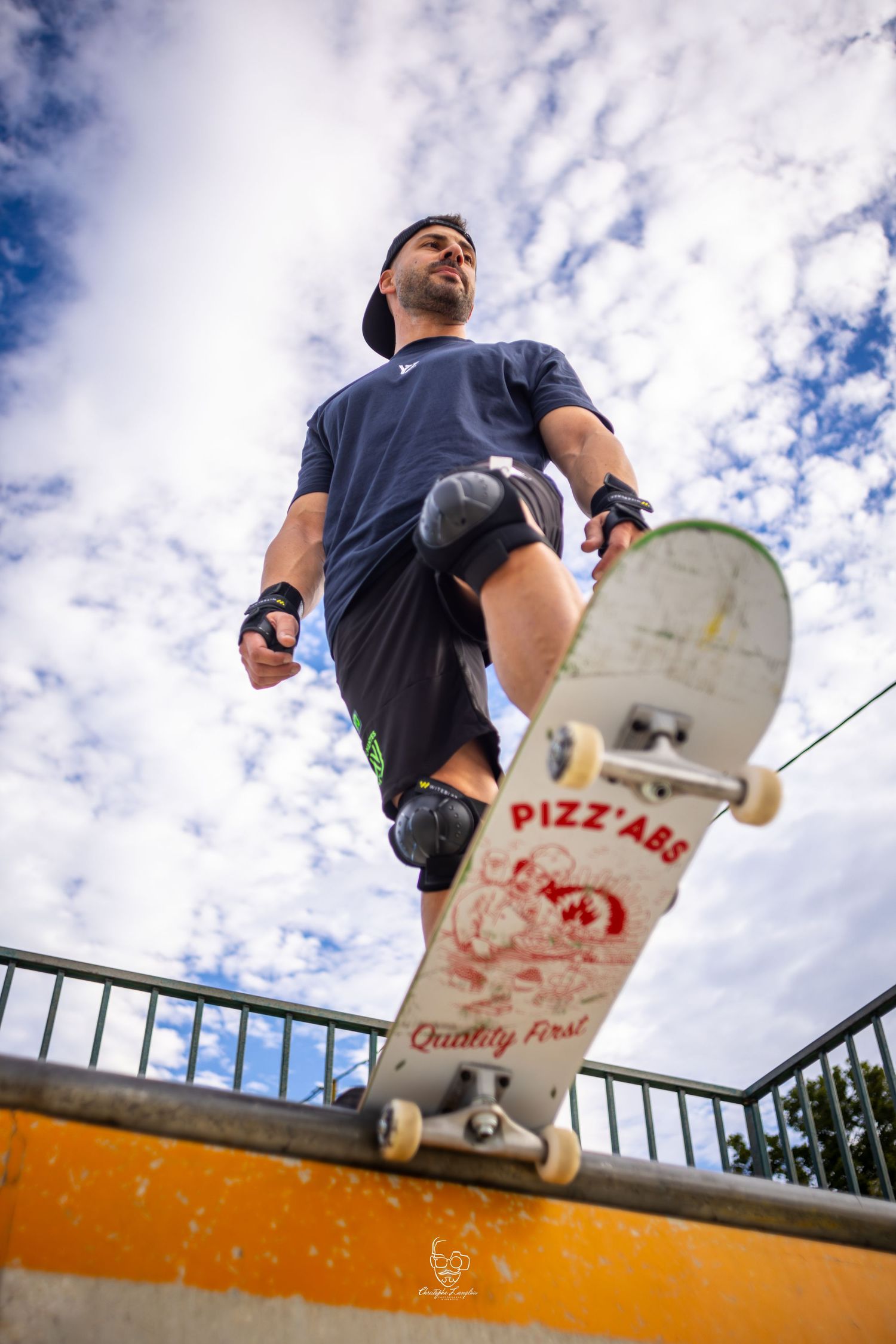 A skateboarder performs a trick sequence on an orange ledge against a cloudy sky with a pizza box design board.