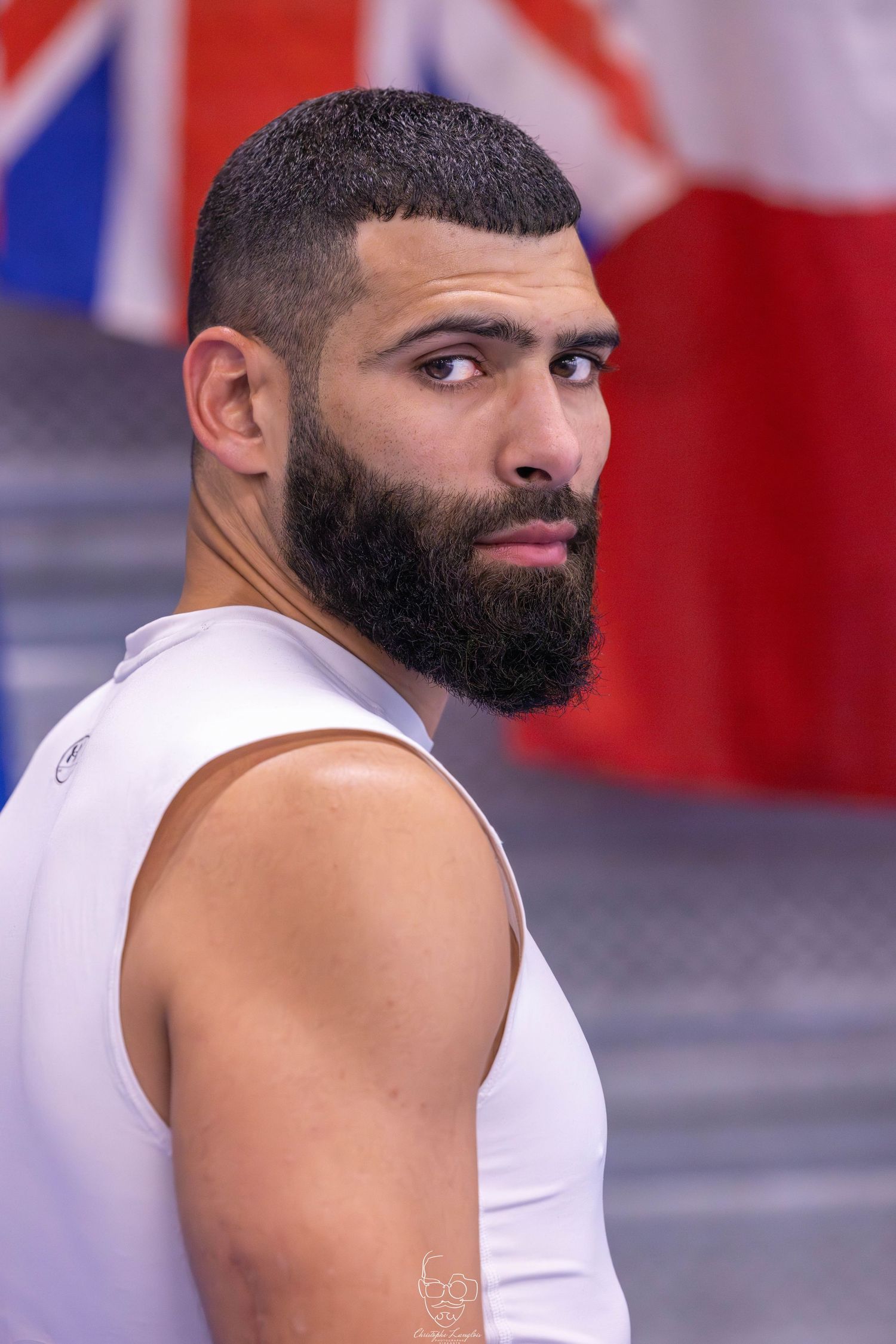 Athletic man in white tank top posing in front of British flag backdrop at sports facility.