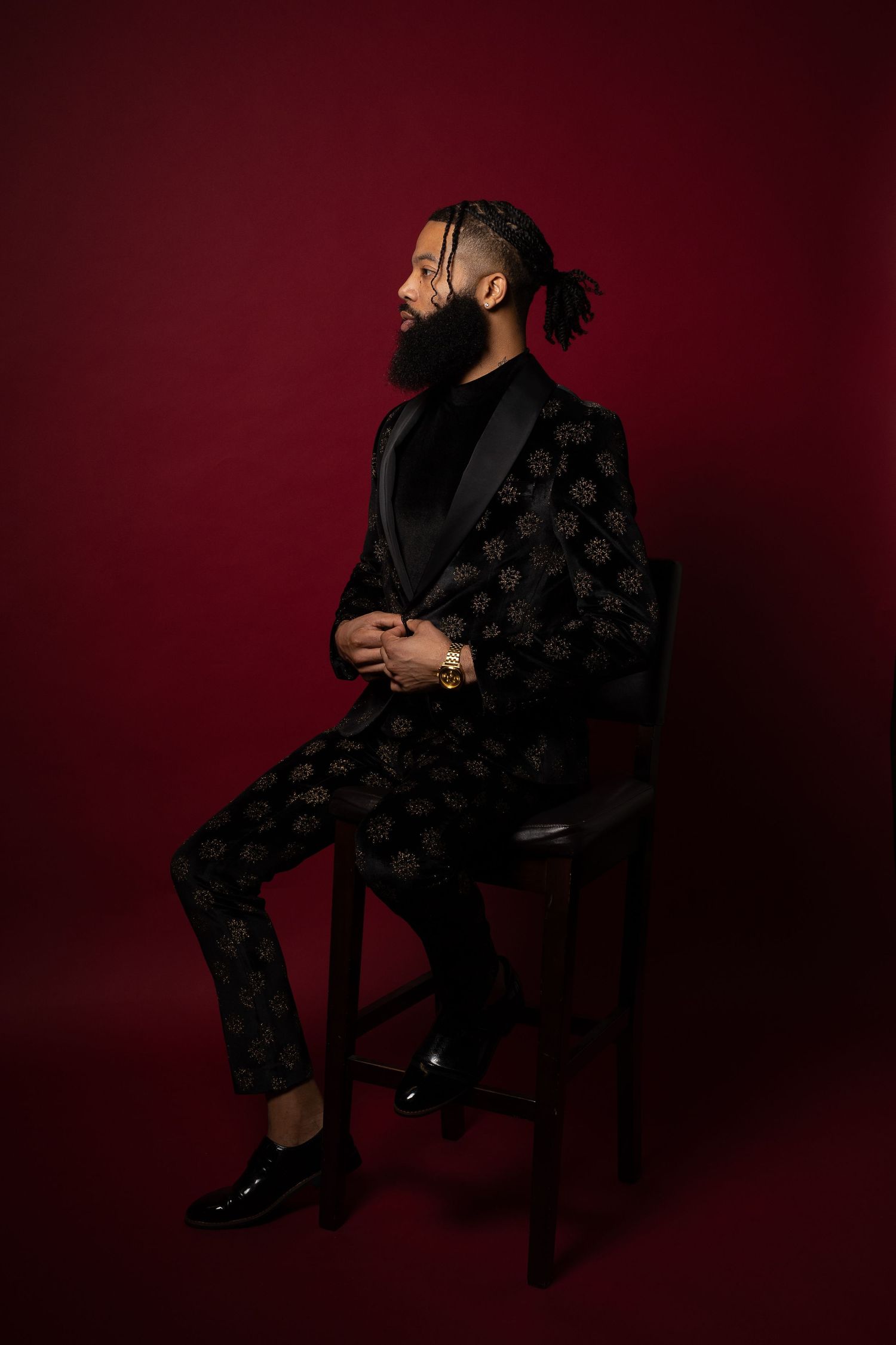 Man in patterned suit sits elegantly on chair against burgundy backdrop in professional photo shoot.