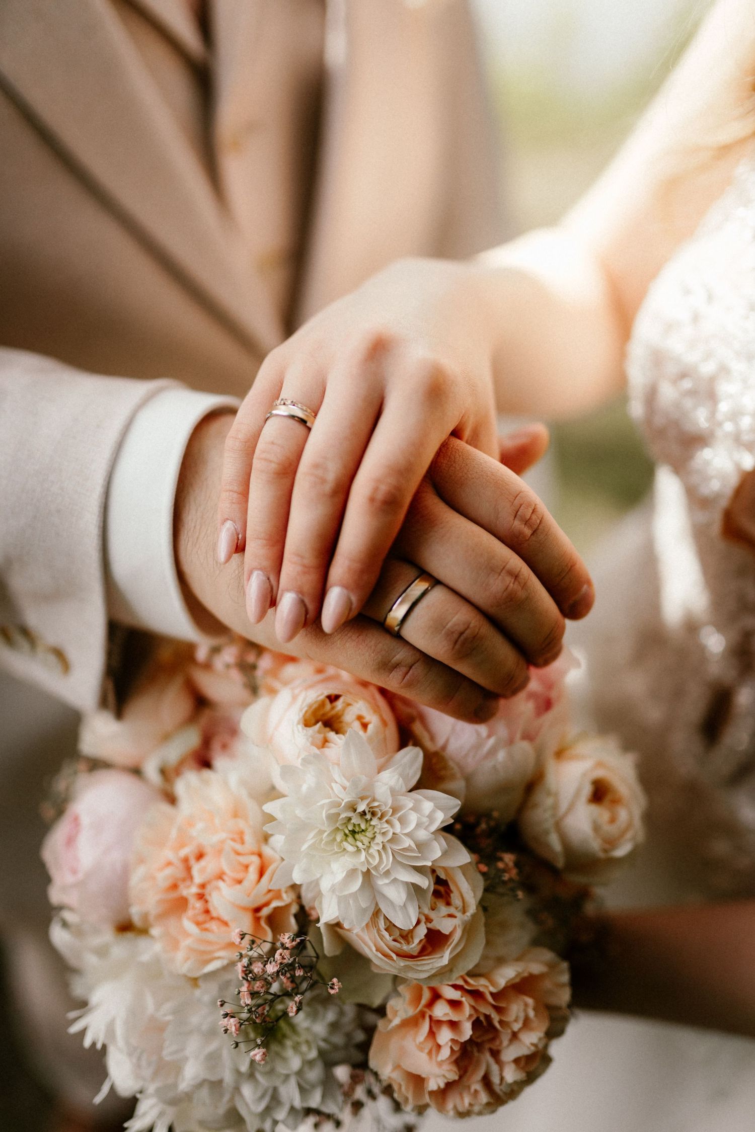 Newly married couple displaying wedding rings while holding a delicate bouquet of peach and white flowers.