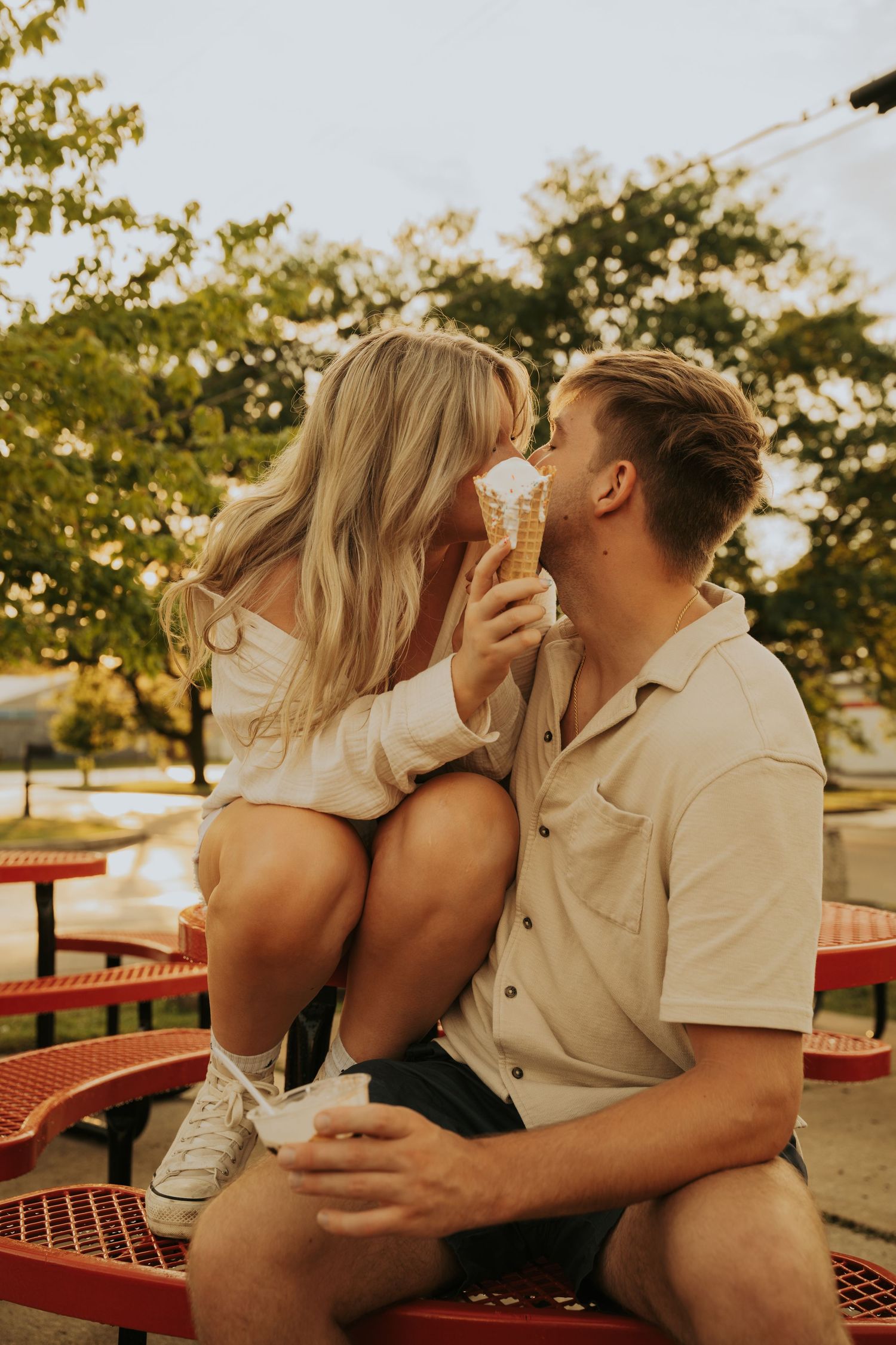 A couple shares ice cream cones while sitting on a red bench during golden hour in a park setting.