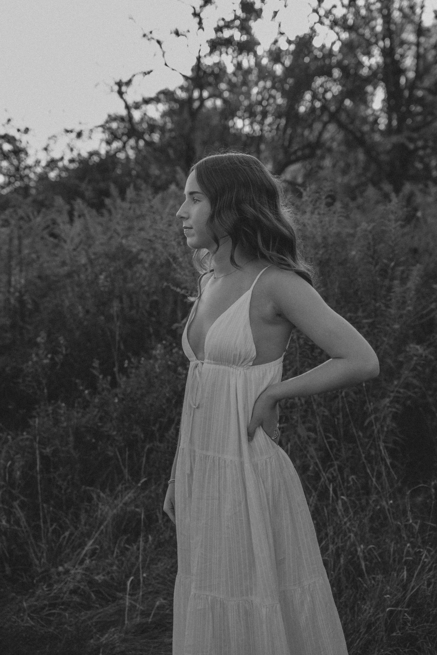 A person in a flowing white sundress poses gracefully in a natural outdoor setting with tall grass and foliage.