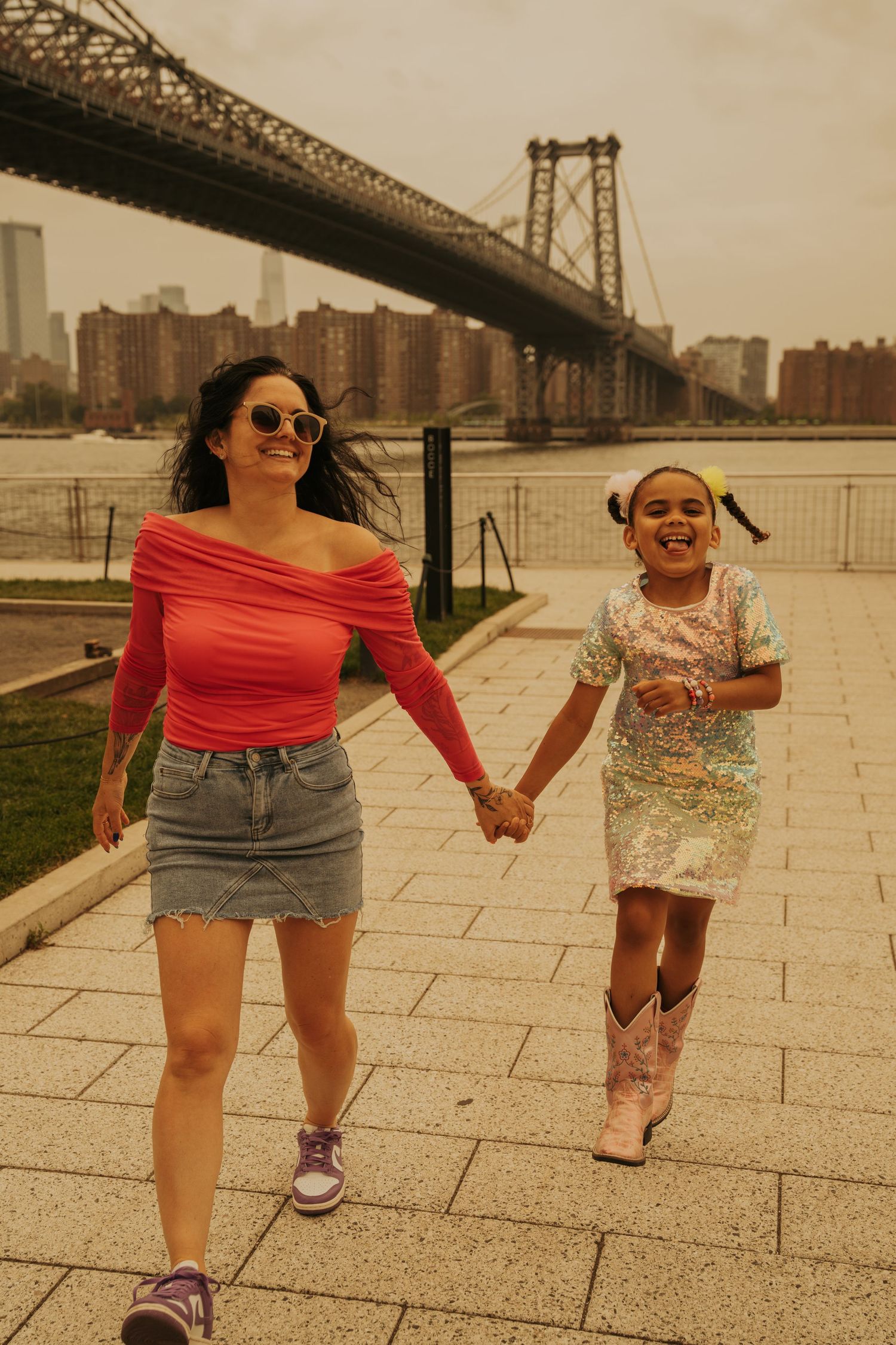 Two people holding hands and walking along the waterfront near the Williamsburg Bridge in New York City on a sunny day.