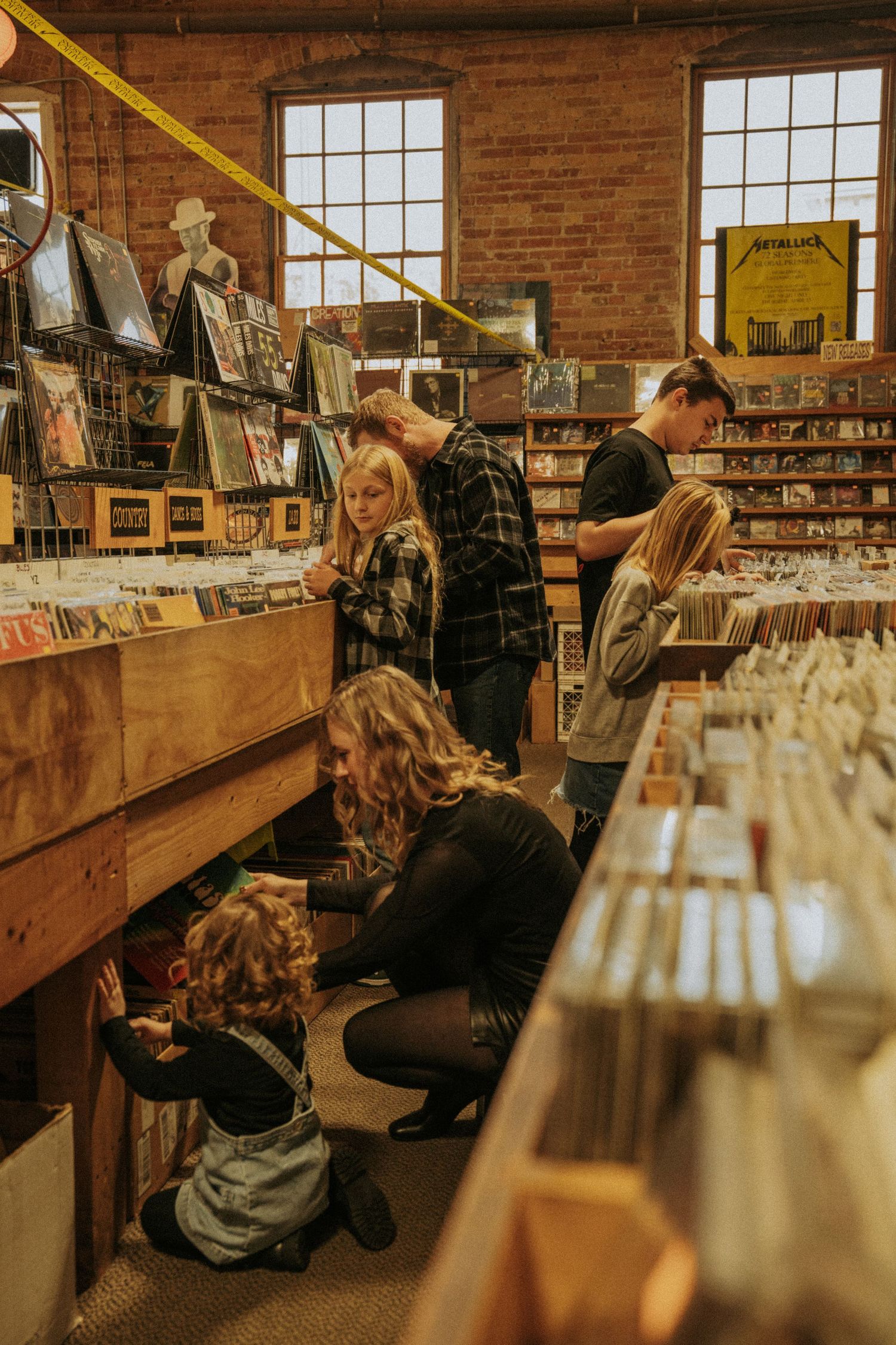 Two people browsing through vinyl records in a rustic brick-walled record store with wooden bins and large windows.