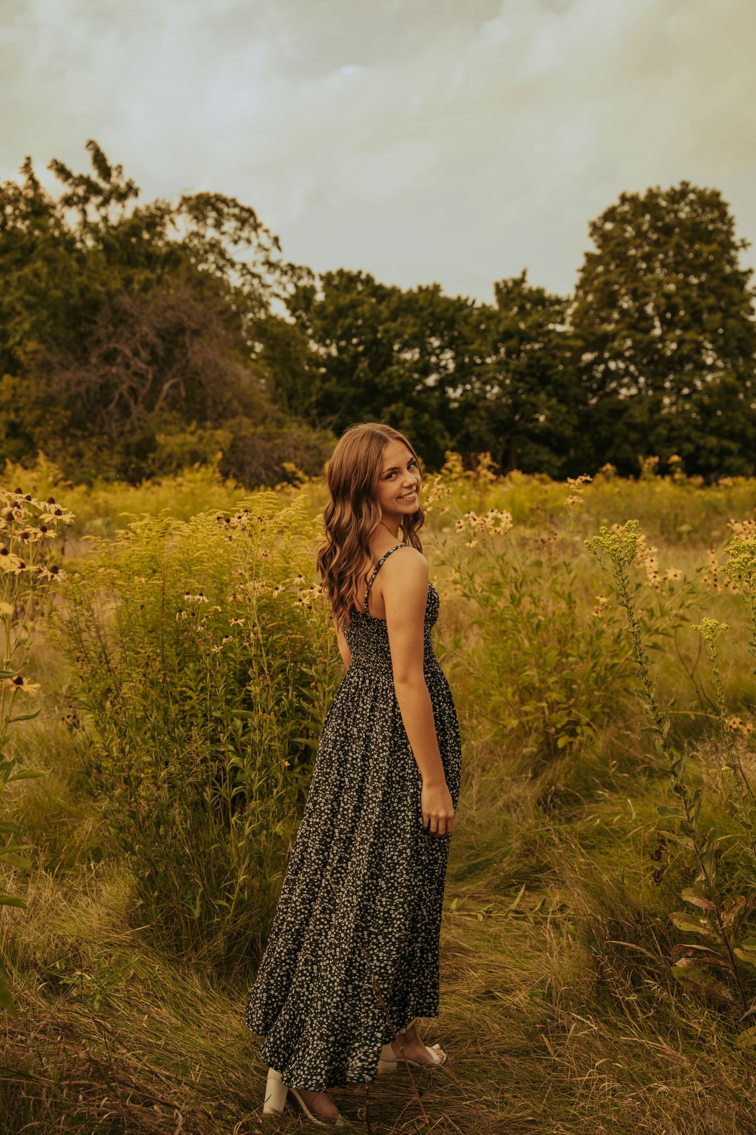 A person in a floral maxi dress stands in a golden meadow at sunset with trees in the background.