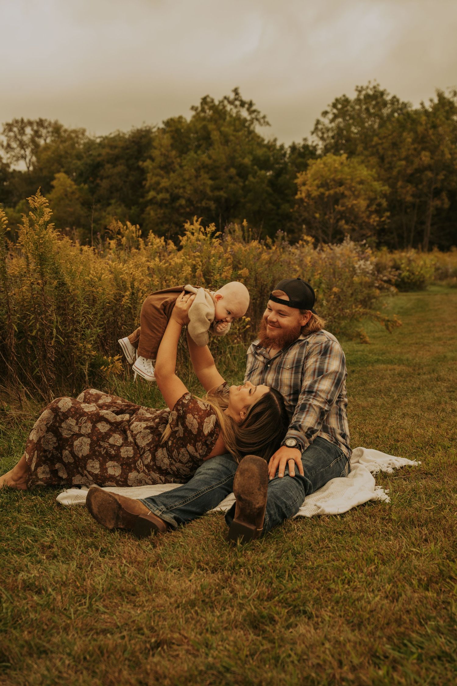 A couple in casual autumn attire lying on grass while sharing intimate moments in a golden-lit field at sunset.