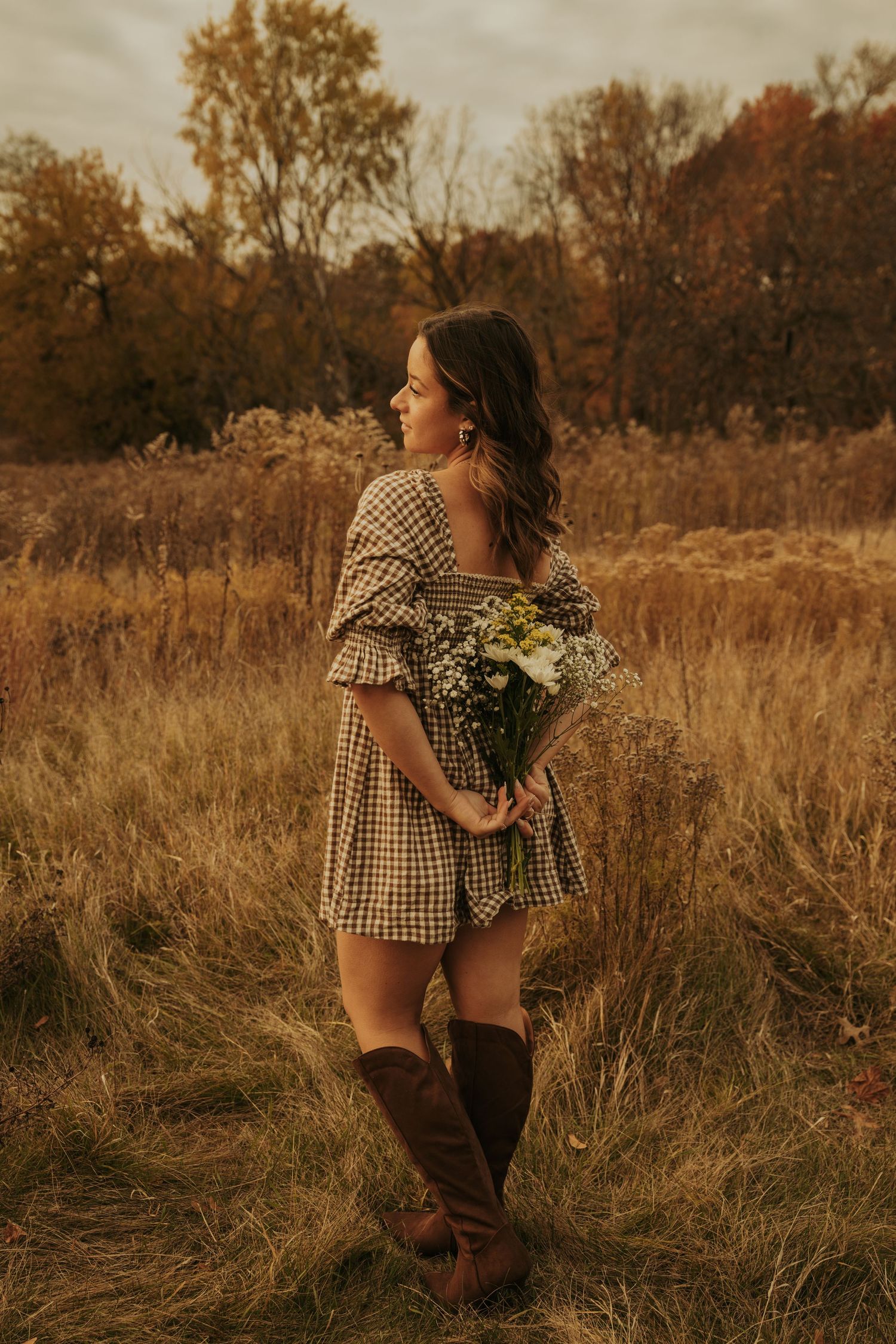A person in a gingham dress holds wildflowers while walking through a golden autumn field at sunset.