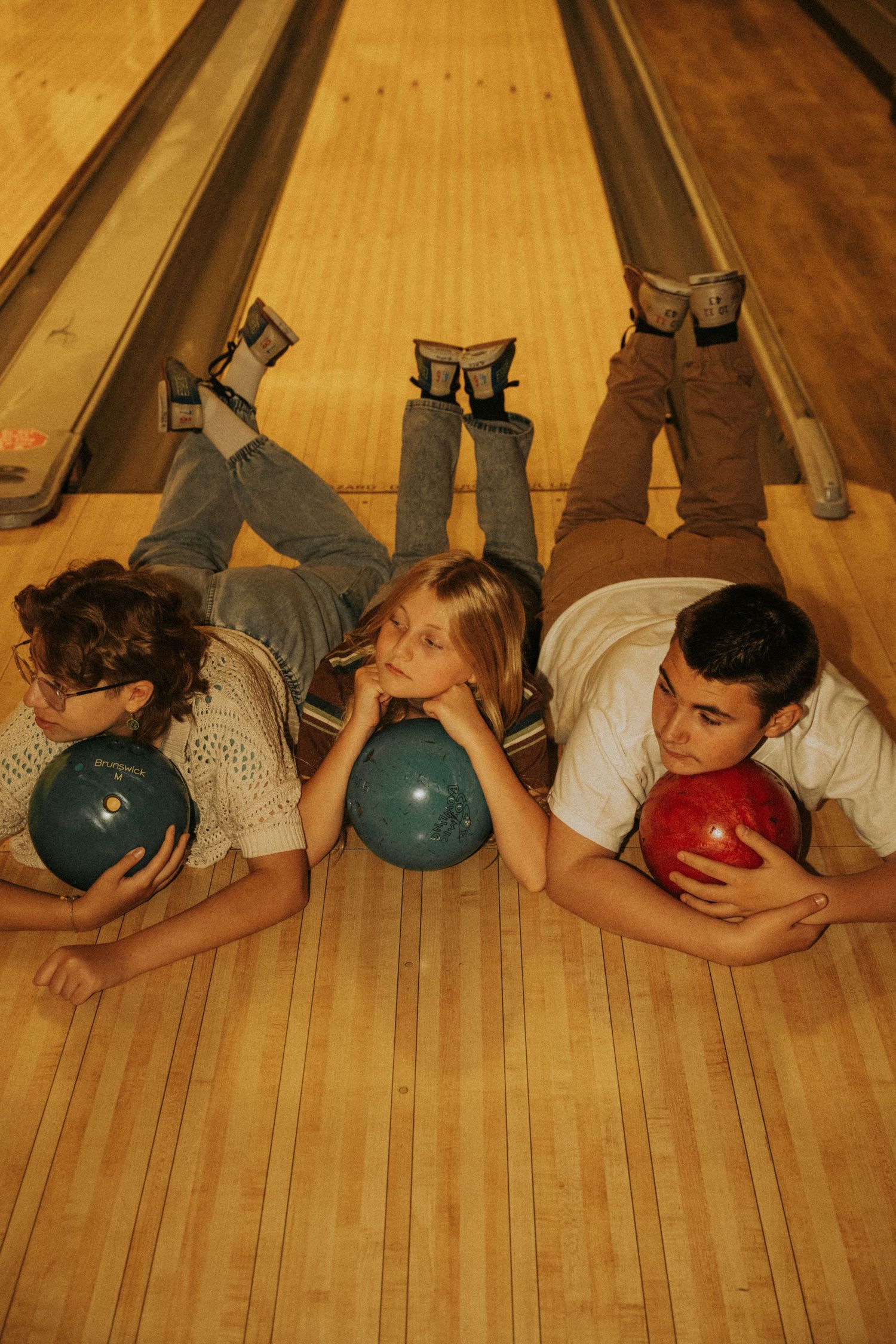 A sequence of bowling balls and players lying face down on wooden lanes at a bowling alley.