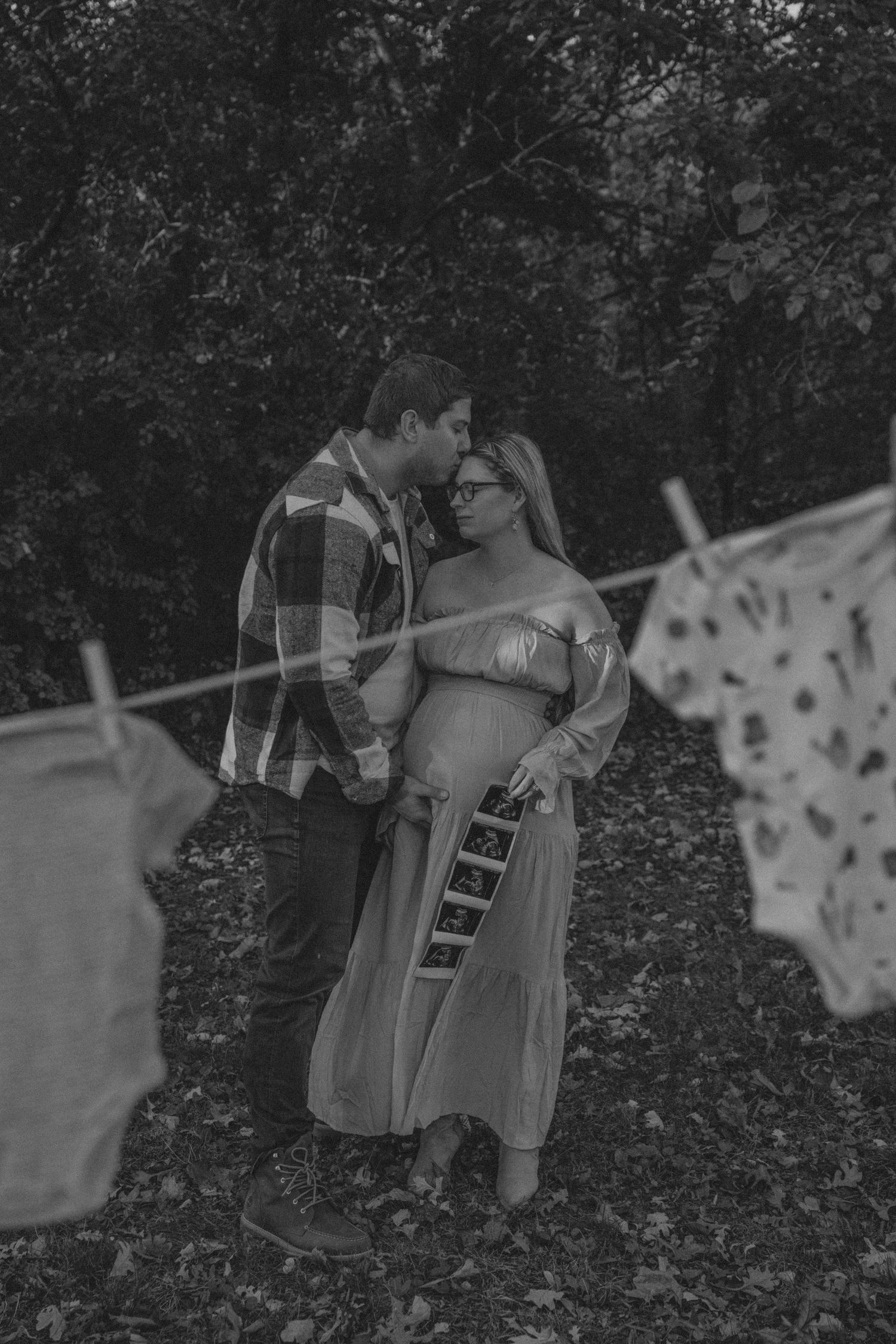 A romantic black and white maternity photo shows a couple embracing by a clothesline in a natural outdoor setting.