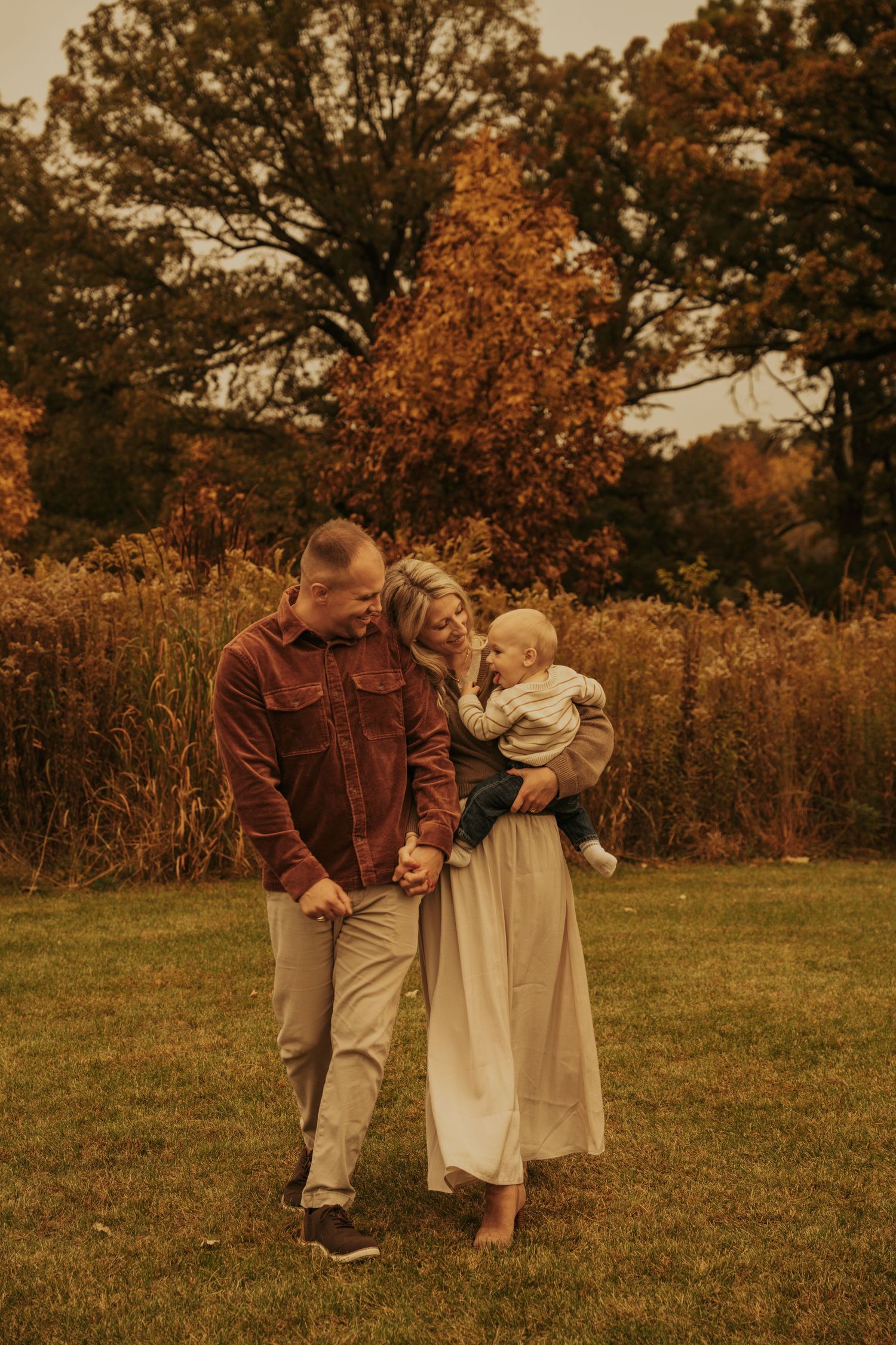 A couple in brown and cream outfits walks together through a fall meadow at sunset creating a romantic autumn scene.