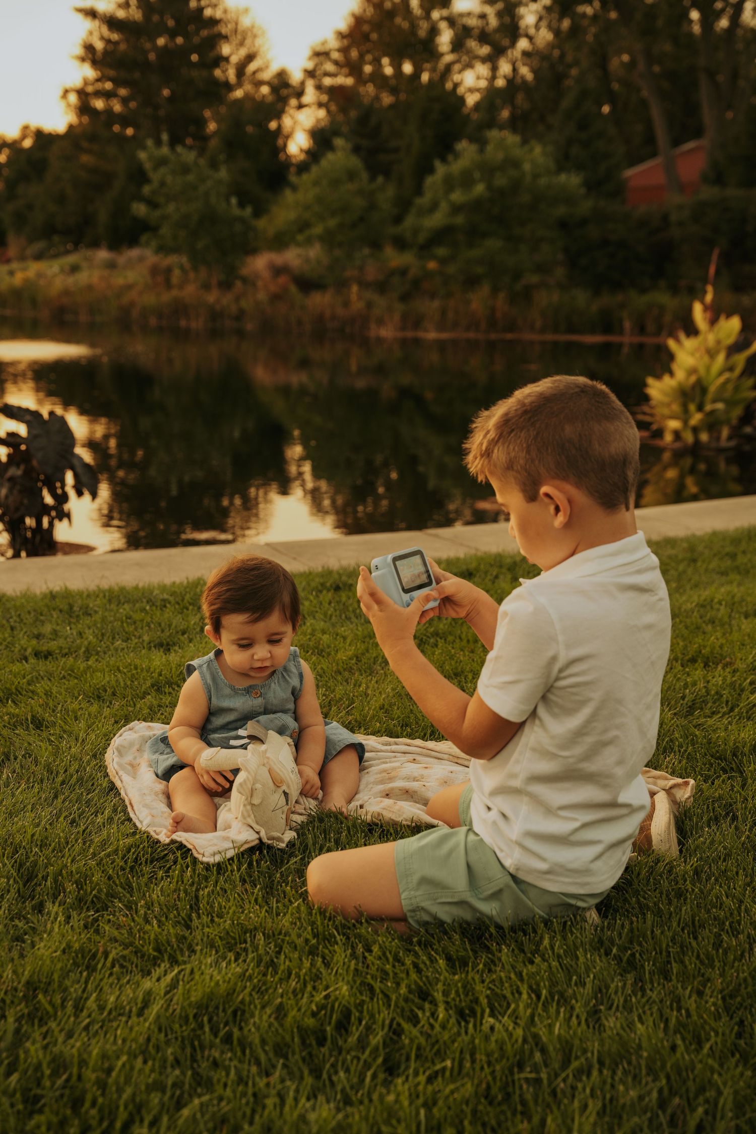 Two children sit on grass by a pond at sunset while one takes a photo with a device.